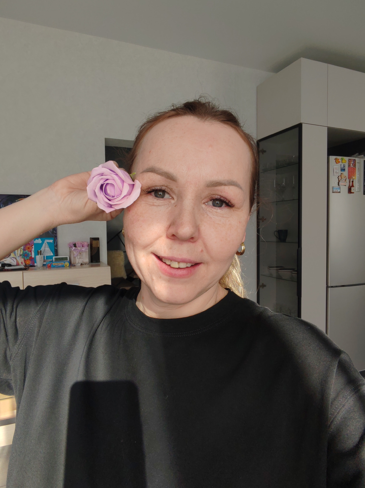 A fair-skinned woman with reddish-blonde hair pulled back poses in a selfie. She is smiling and holding a light purple rose near her left eye. She wears a black crew neck shirt and gold hoop earrings. The background includes a white kitchen with cabinets and a refrigerator. Other objects visible include various items on a counter or shelf behind her. The location is implied to be Chaikovsky, Russia.