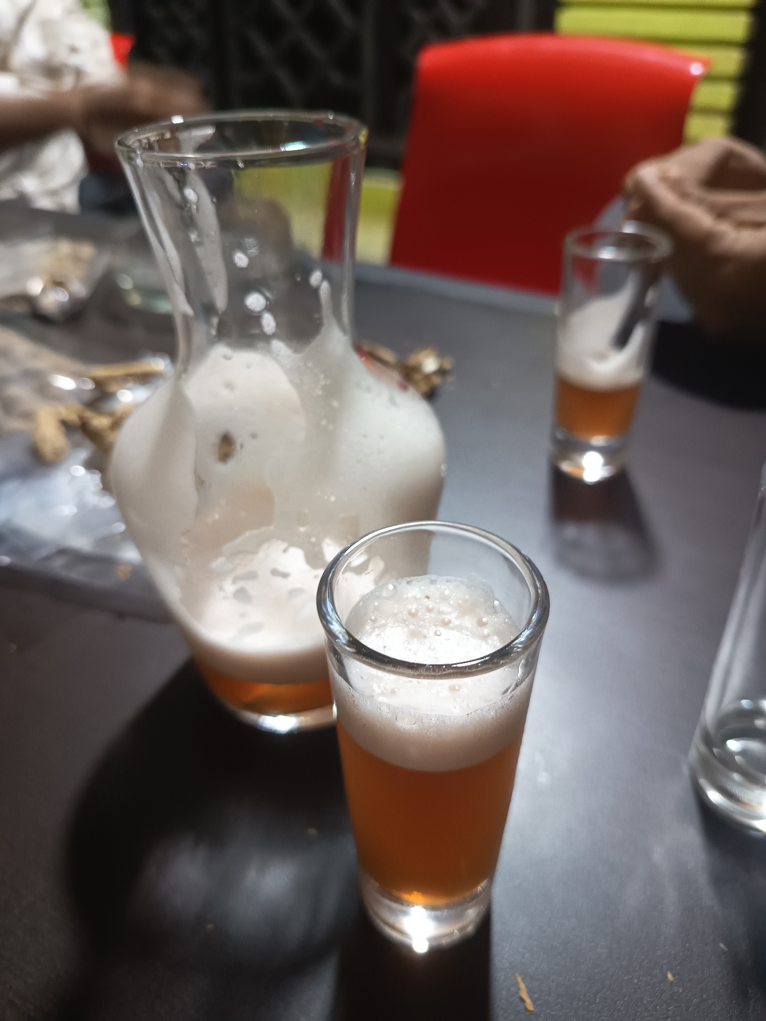 A decanter filled with foamy, amber liquid is placed on a dark, textured table. Beside it are three small shot glasses, two of which are filled with the same foamy, amber liquid. The decanter is made of clear glass and has a wide base that tapers to a narrower neck. The liquid inside is frothy, with a thick layer of foam on top. The shot glasses are also made of clear glass and have a similar amber-colored liquid with a foamy head. In the background, a red chair is visible, along with blurred figures of people. The scene appears to be indoors, possibly in a restaurant or bar. The location is Tapak Tuan, Indonesia.