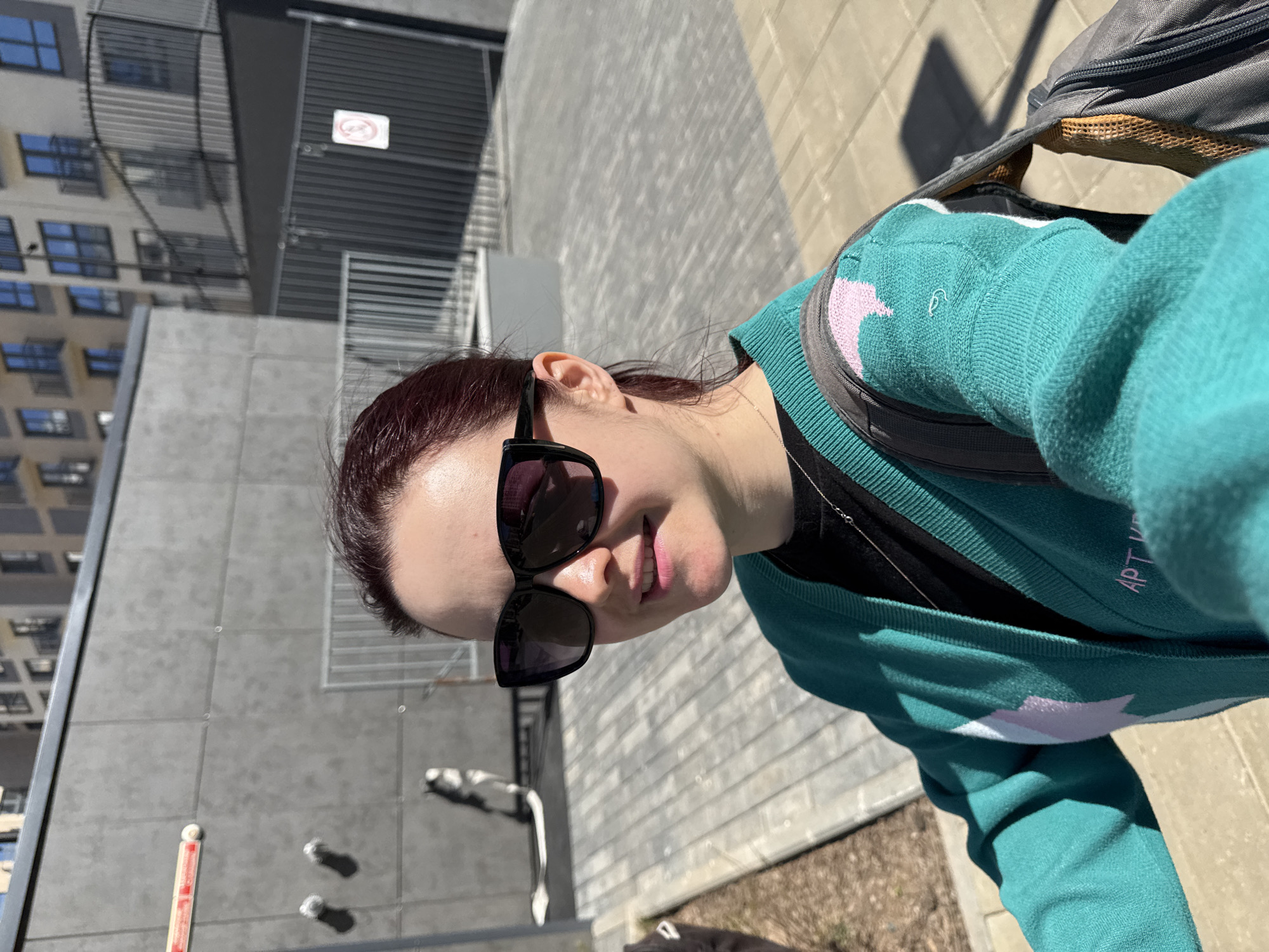 A woman with dark red hair, wearing a turquoise sweater with light pink and white stars, and black sunglasses, is standing on a paved sidewalk. She is smiling slightly. Part of a building is visible behind her, and a sign is on the building. A backpack is visible on her back. The location is likely Izhevsk, Russia.