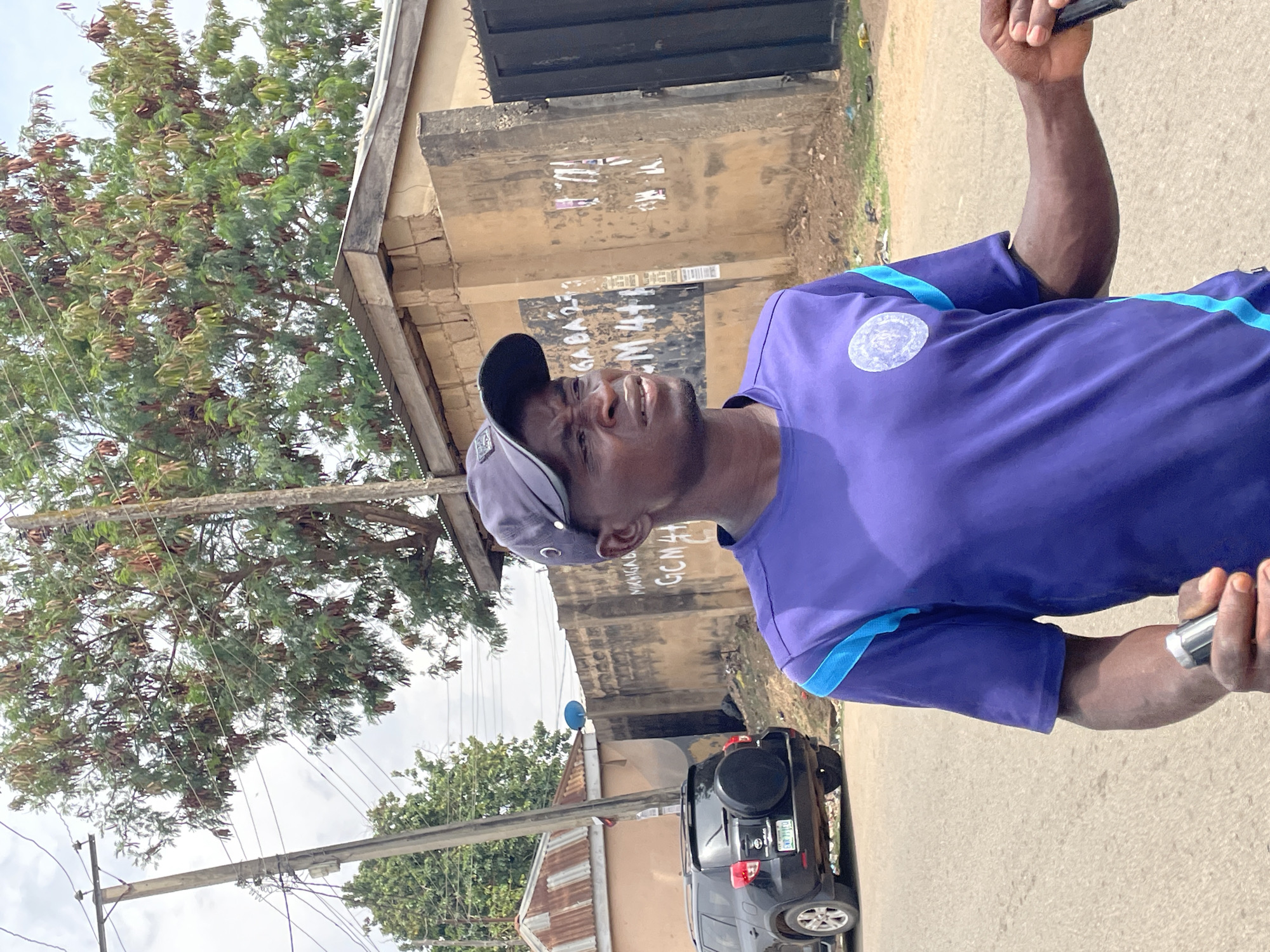 The image depicts a man standing on a street in Jos, Nigeria. He is wearing a purple shirt with blue trim and a grey cap. He is looking upwards and holding an object in his right hand. Behind him is a street with buildings, a tree, and a black SUV. One building has faded writing. The environment seems urban and somewhat congested.