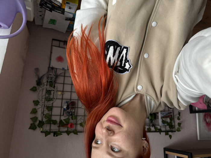 A woman with vibrant orange hair is angled upside down. She is wearing a beige varsity jacket with a black and white "MA" patch. Her fair skin is visible with a lip ring and multiple earrings. The background includes a pink wall, a black wire grid decorated with faux ivy and a dreamcatcher, a purple watering can, and framed artwork. The overall impression is casual and youthful.