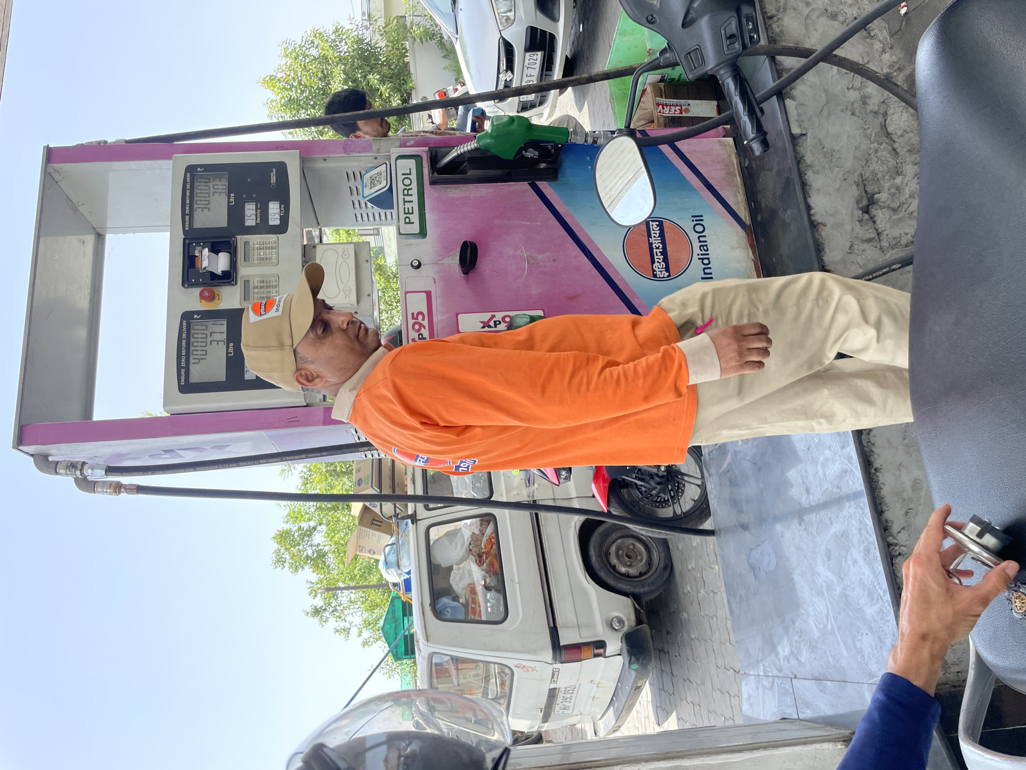 The image shows a man in an orange shirt and khaki pants standing at a gas pump at an Indian Oil petrol station. The pump is pink and blue with the "Indian Oil" logo prominently displayed. The pump display shows "400.00" and "3.71 Litre" and "300.00" and "3.03 Litre". The gas nozzle is green. Behind the man, there is a white Omni van with the license plate "RJ 39 U092". A black motorbike is parked behind the van. In the background, a white car with the license plate "RJ 09 F 7029" can be seen. The location is Sidhpur, India.