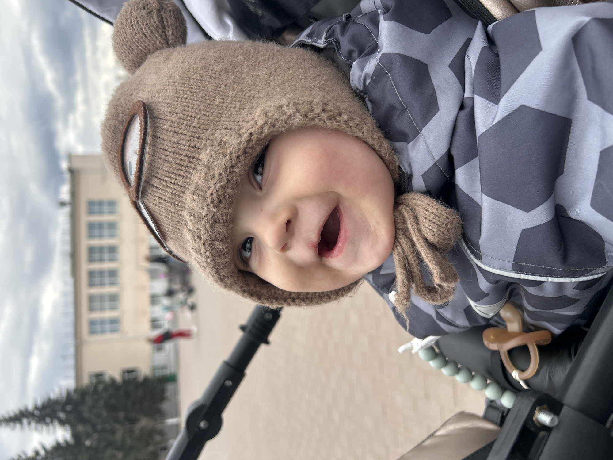 A close-up, rotated shot captures a joyful baby from a low angle, looking upwards and smiling with an open mouth. The baby is wearing a light brown, knitted hat with bear-like ears and decorative brown straps resembling goggles on the sides. Their eyes are wide and bright. They are dressed in a warm, padded jacket with a grey and dark grey geometric, possibly hexagonal, pattern.
The baby appears to be in a stroller, parts of which are visible in the foreground, including a black frame, a light green beaded accessory, and a brown pacifier hanging from the side. In the blurry background, a large building with multiple windows is visible, suggesting an urban or suburban setting. The sky above is overcast with clouds. The natural lighting indicates it is daytime. The overall mood is cheerful, reflecting the baby's happy expression. There is no visible text in the image. The scene is set in Chaikovsky, Russia.