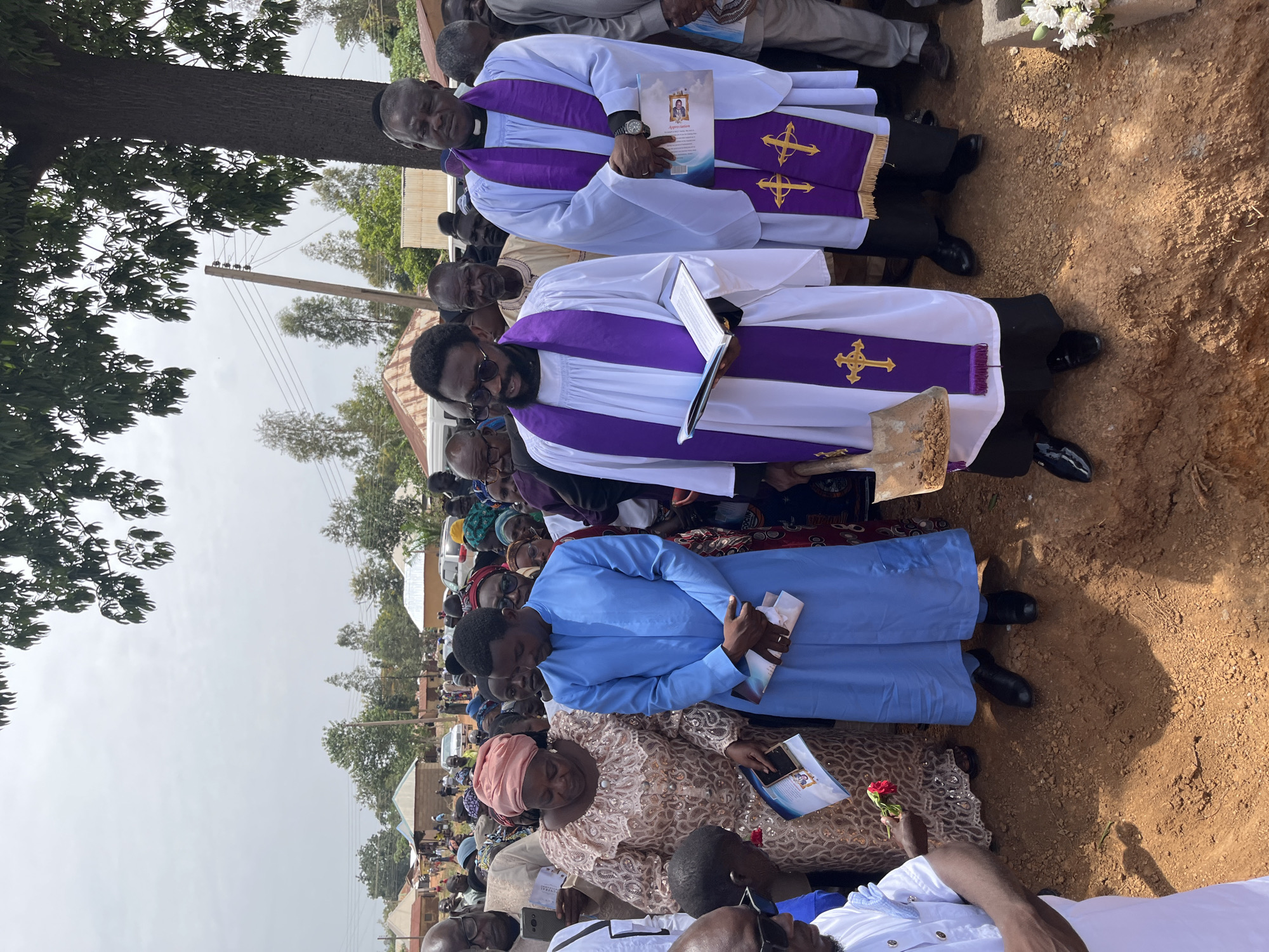A solemn outdoor gathering is underway in Dogon Dinia, Nigeria. Two men, possibly clergy, stand in white robes with purple stoles marked with gold crosses, each holding a book or document. Numerous attendees, including women in patterned dresses and headwraps and men in a mix of clothing, surround them. One woman holds a red rose. The setting appears to be a gravesite, evidenced by the dirt ground, and a possible headstone with white flowers is visible in the background. There is an electric utility pole. The sky is overcast.