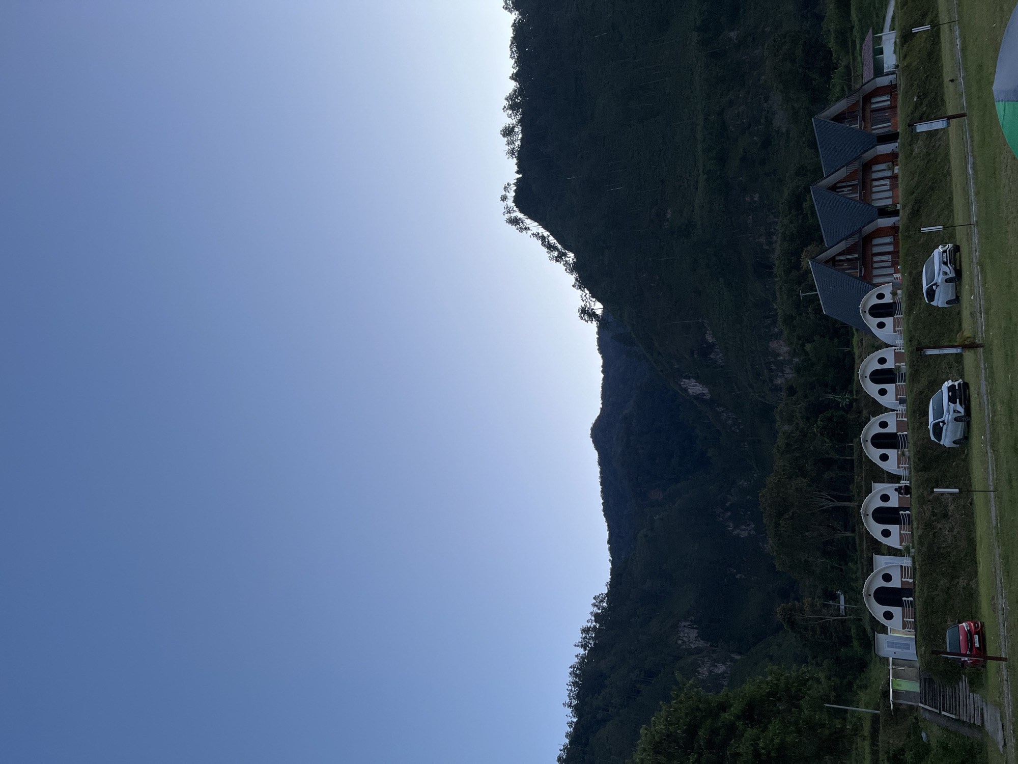 The image depicts a serene evening scene in Merodot, Indonesia. The sky is a clear, gradient blue, transitioning to a lighter hue towards the horizon. In the foreground, a row of unique dome-shaped cabins and three triangular A-frame houses are situated on a grassy slope. A red car is parked to the left of the cabins, and a white SUV is parked in front of the A-frame houses. The middle ground is characterized by lush green grass and several trees. The background is dominated by a majestic, dark green mountain range covered in dense forestation. The mountain slopes are steep, with scattered trees silhouetted against the sky along the ridgeline. The overall atmosphere is peaceful and tranquil, suggesting a secluded natural retreat.