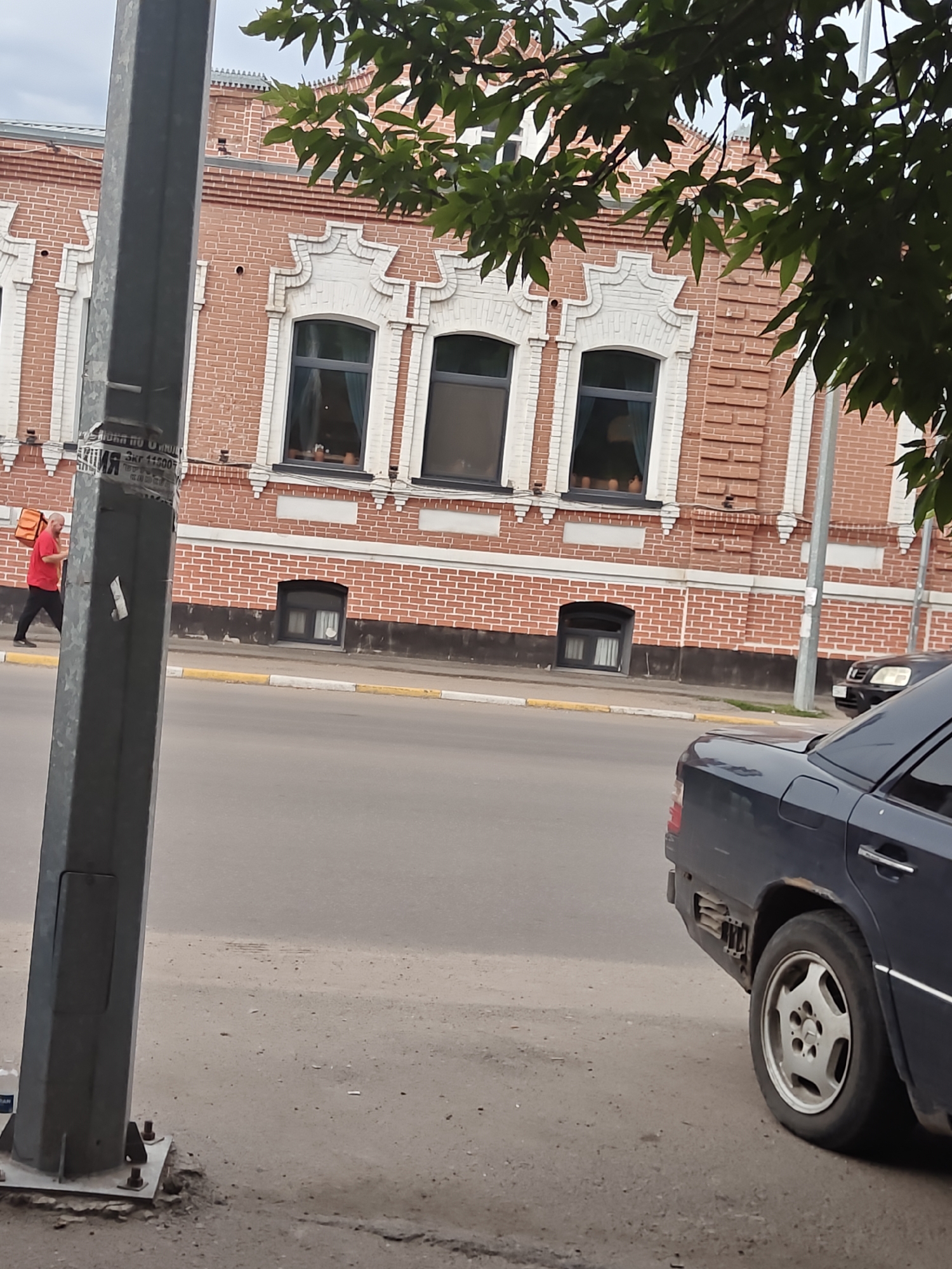 The image shows a street scene in Petropavl, Kazakhstan, featuring a brick building with decorative white accents and several windows. A man in a red shirt and black pants is walking on the sidewalk, carrying an orange bag. A dark blue sedan is parked on the right side of the street, partially obscuring the lower portion of the building. A lamppost stands in the foreground on the left, with some peeling stickers on it. Trees with green leaves are visible overhead, partially covering the top of the building. The ground is paved, with a yellow line marking the edge of the sidewalk. The sky is overcast.