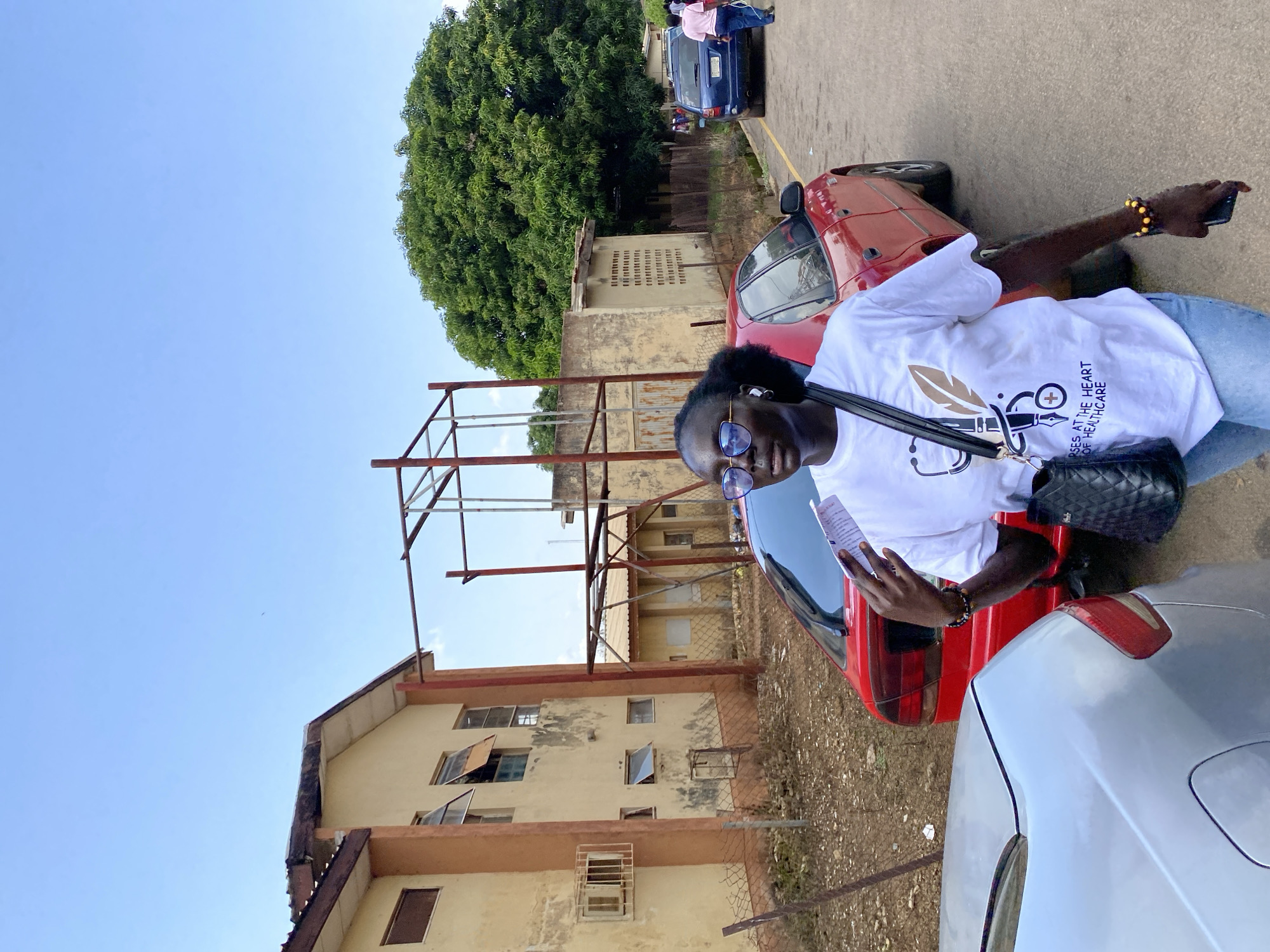 The image depicts an outdoor scene in Jos, Nigeria, with a woman in the foreground holding a pamphlet. She is wearing a white t-shirt with a stethoscope and feather graphic and the text "NURSES AT THE HEART OF HEALTHCARE," blue jeans, and round sunglasses. She carries a black quilted handbag over her shoulder and wears beaded bracelets on her wrist. Behind her, a red car is parked, and further down the road, a blue SUV and two people in pink shirts are visible. The background features a two-story building with cream-colored walls and orange trim, several open windows, and a chain-link fence. A large, leafy green tree is prominent to the right of the frame, and the sky is clear and blue. The ground is an asphalt road with some dirt and debris visible along the roadside. The general setting suggests a modest urban or semi-urban environment.