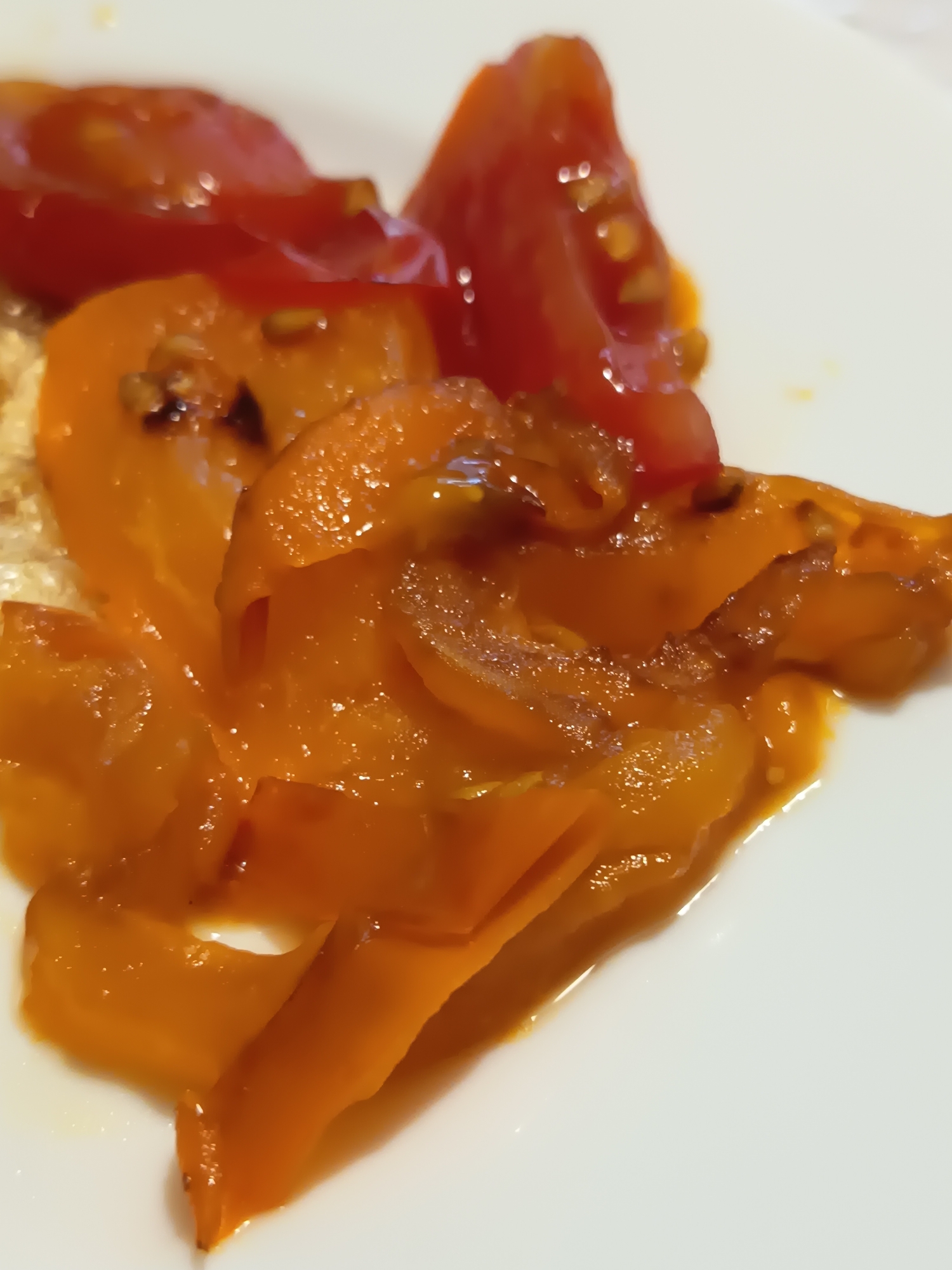 A close-up, slightly out-of-focus shot captures cooked vegetables, primarily sliced orange bell peppers and some red tomatoes, arranged on a white plate. The vegetables appear soft and slightly glossy, suggesting they have been sautéed or roasted. The lighting creates highlights on the glossy surfaces, and some seeds are visible within the peppers. The background is a plain white surface, likely a table or countertop. There is no indication of the location being Izmail, Ukraine.