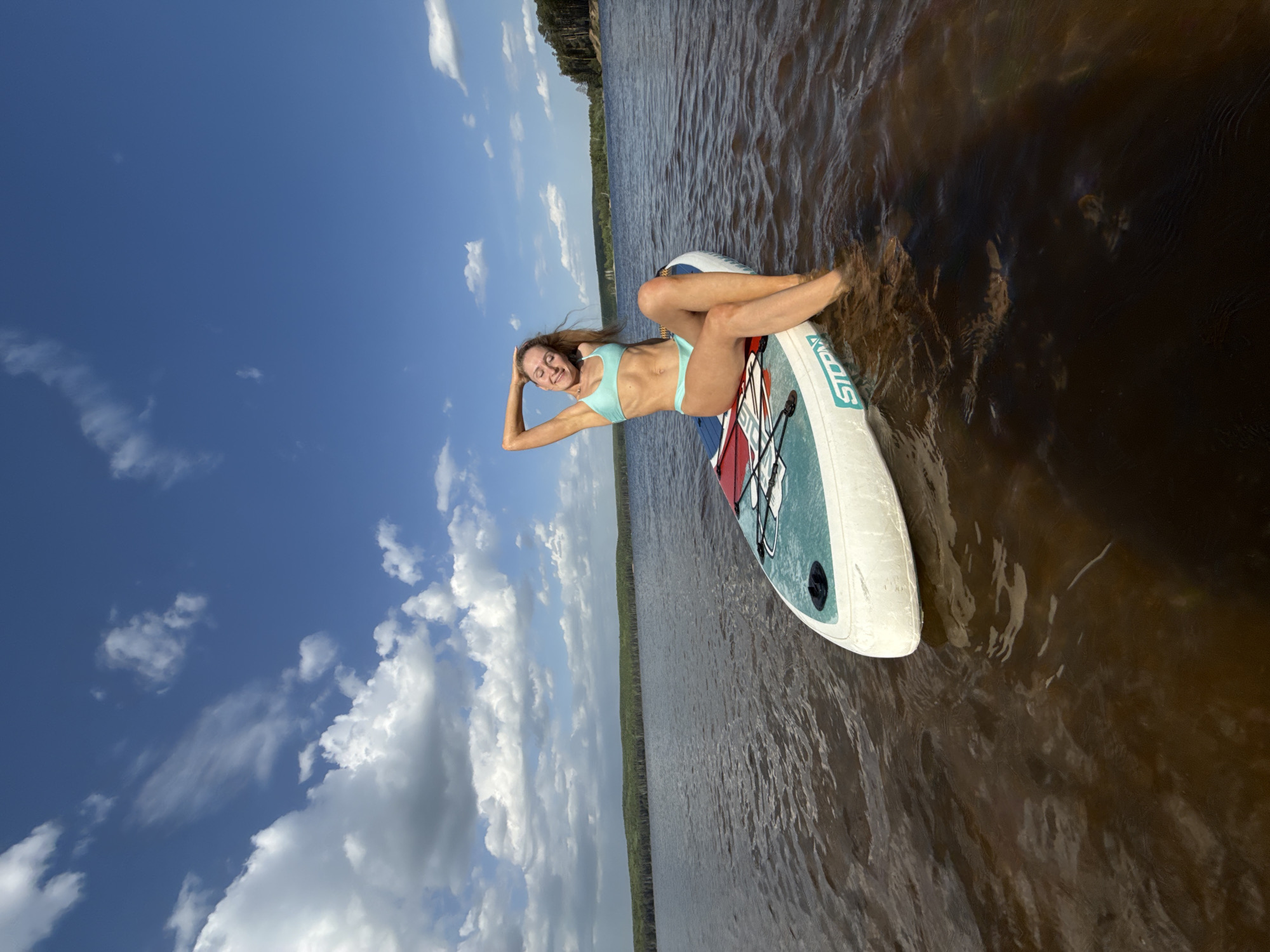 A woman is reclining on a paddleboard in a body of water. She is wearing a light blue bikini and has her hands behind her head. The paddleboard is mostly white with red and blue accents. The water appears to be brown or dark, and the sky is blue with white clouds. Trees are visible along the shoreline in the distance. The image was taken near the city of Chaikovsky, Russia, based on the background landscape and typical water body in the area.
