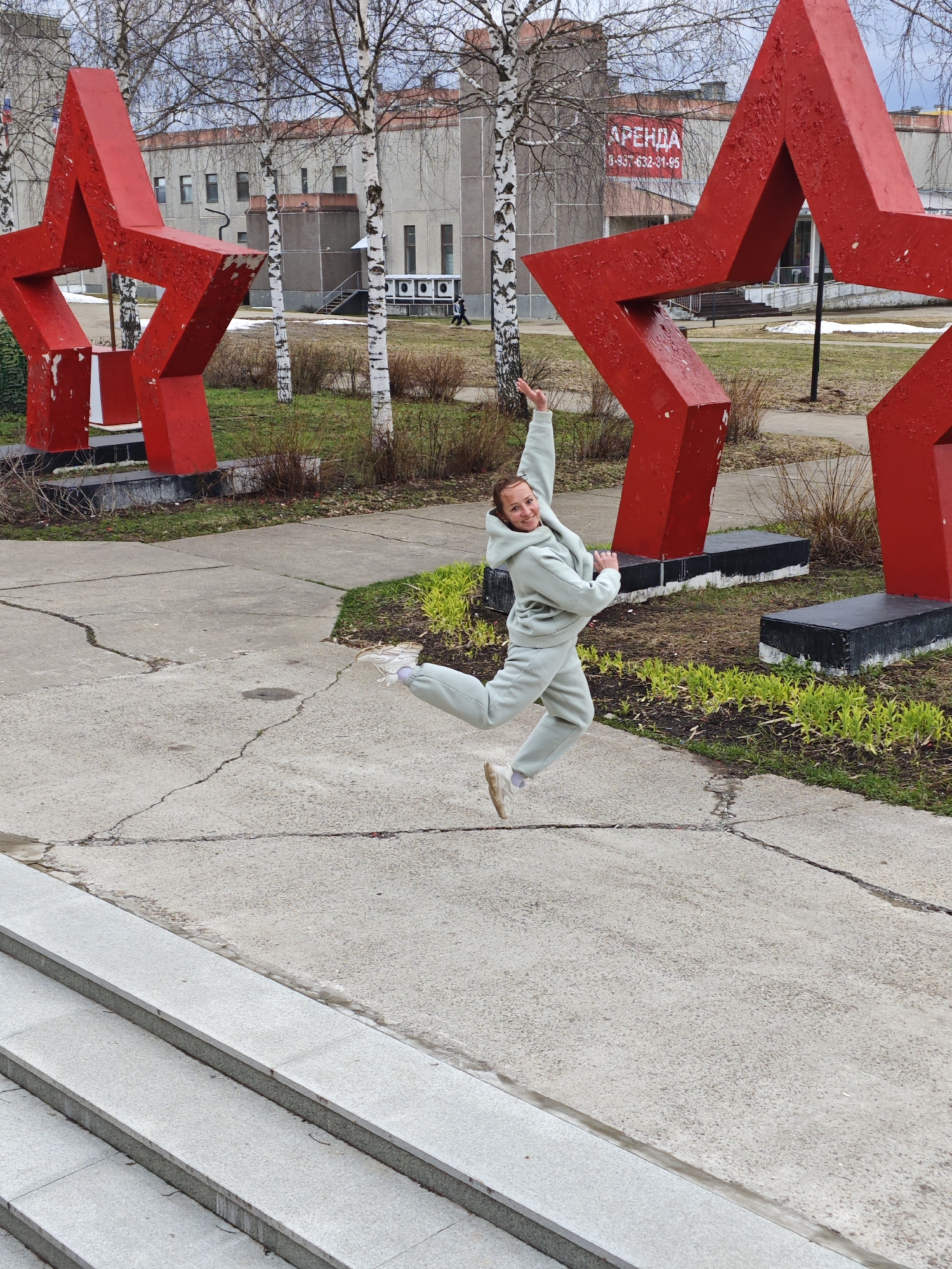 A woman in a light green sweatsuit jumps in the air between two large, red, five-pointed star sculptures. The location is Markovskii, Russia. The sculptures are placed on grass and concrete. The background includes bare trees, a multi-story building, and a sign in Russian on the building advertising “APEHDA,” presumably a rental. The foreground features gray concrete steps. The woman is smiling and has one arm raised.