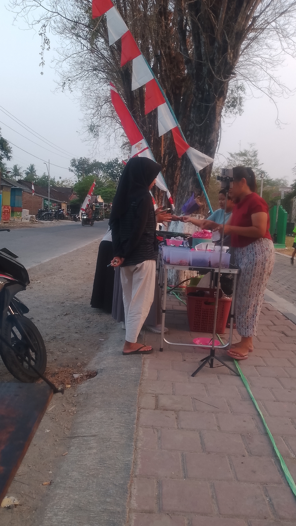 The image depicts an outdoor scene in Karanglangu, Indonesia. In the foreground, a motorcycle is partially visible on the left. A paved walkway is to the right, with a dirt and gravel area to the left. A woman wearing a hijab and light-colored pants stands on the walkway, facing a vendor. The vendor, wearing a red shirt and patterned pants, is behind a portable table displaying various food items in clear plastic containers. Another person, partially obscured, stands behind the vendor. Indonesian flags are displayed on poles behind the vendor. In the background, a road runs alongside the walkway with several motorcycles parked along it and buildings on the other side. Trees line the area, with one large tree prominently featured with bare branches. The sky is overcast.