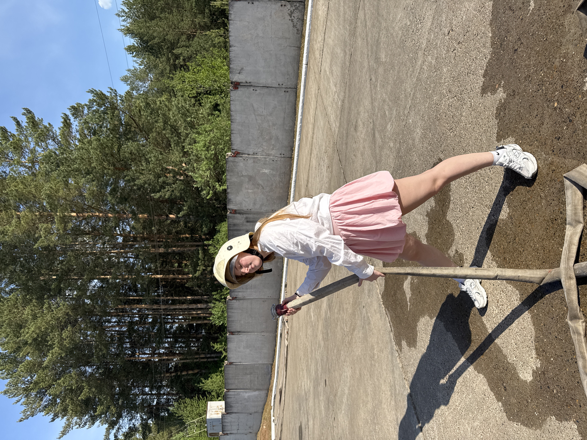 A young person, likely a female, stands outdoors in what appears to be a paved area. The location is unspecified, and there is no confirmation of it being in Rusalevka, Russia. She is wearing a light-colored helmet, a white long-sleeved shirt, and a pink skirt. Her footwear consists of white sneakers. She is holding a fire hose with a red nozzle attachment. Behind her is a concrete wall, and in the background are trees. The weather appears to be sunny, casting shadows.