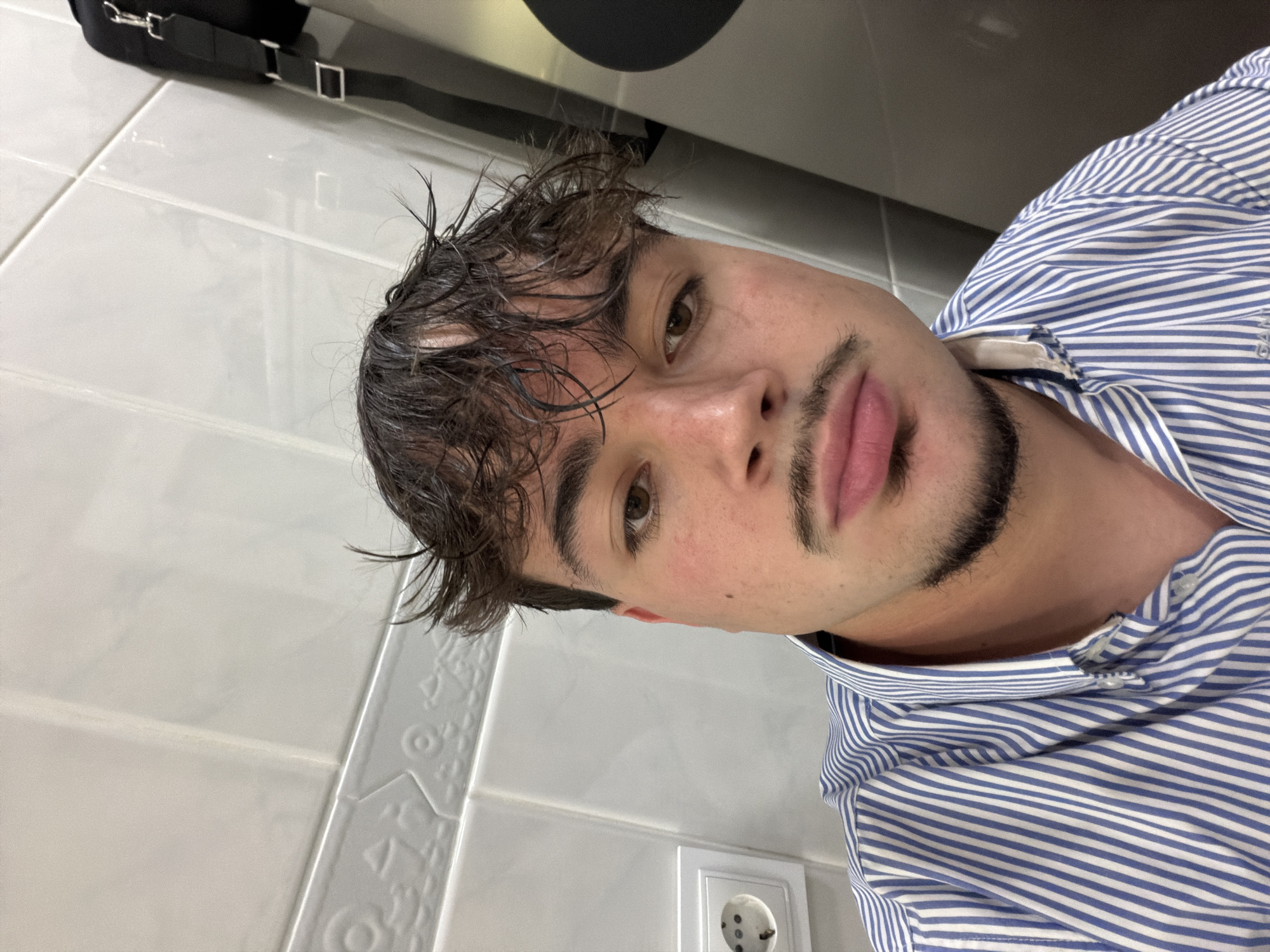A selfie captures a young man in a blue and white striped shirt. His dark, wet hair is styled with a casual, slightly messy look. He has brown eyes and a trimmed beard.
The background consists of white tiled walls, with an electrical outlet visible. A portion of a black bag with a strap is visible in the upper left corner.
While it is possible the image was taken in a bathroom, information regarding the location in Vila Real, Portugal is not present in the image.