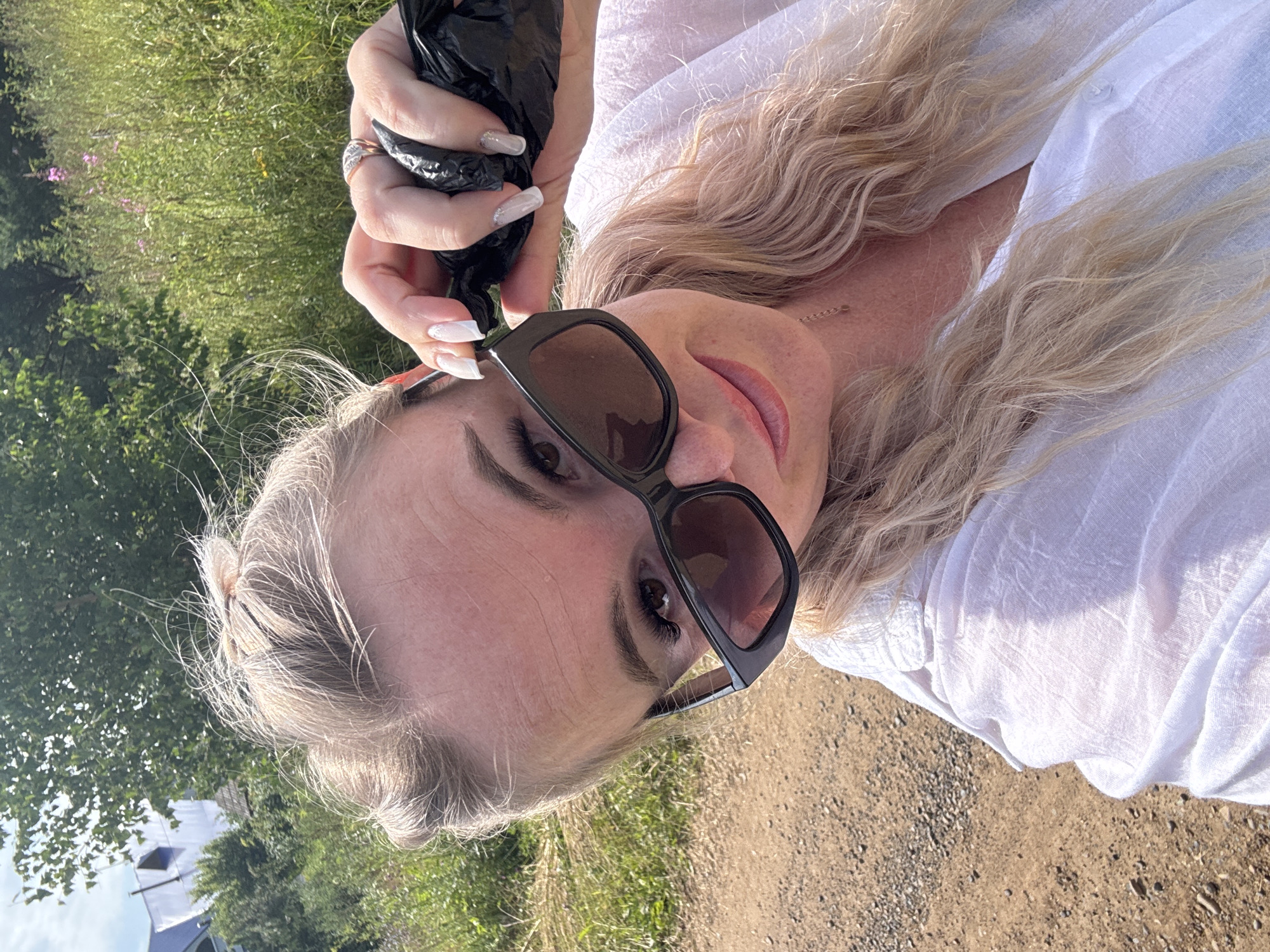A woman with blonde hair pulled up into a bun and loose waves is taking a selfie outdoors. She is wearing large black sunglasses and a white shirt. Her right hand is raised, holding a black plastic bag. The background shows greenery and a dirt path. The location is identified as Pozdyshki, Russia.