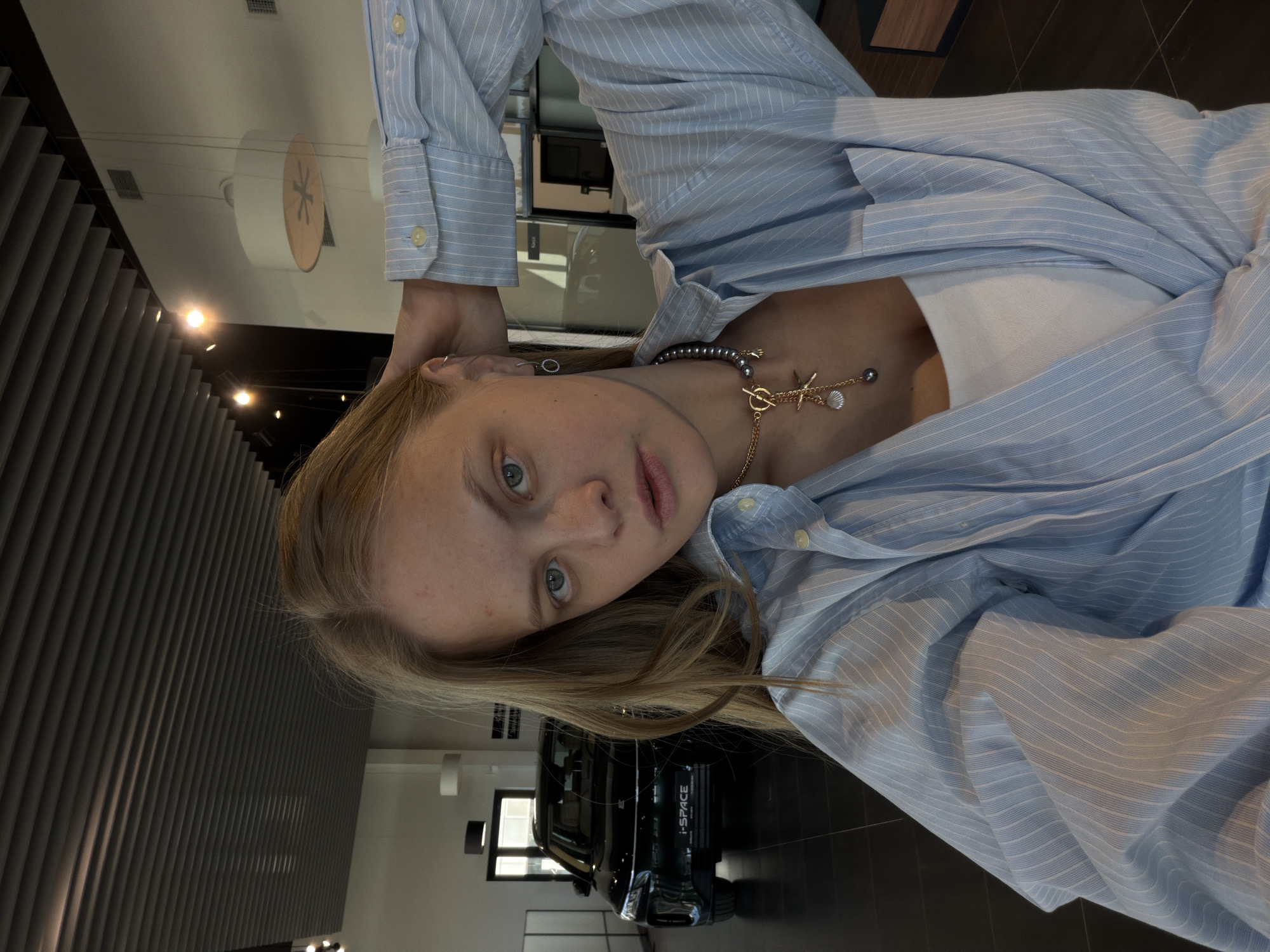 The image shows a blonde woman with fair skin and blue eyes wearing a light blue striped shirt and a necklace with a starfish pendant. She is in what appears to be a car dealership in Izhevsk, Russia, as indicated by the "I-Space" logo on a dark-colored vehicle in the background. The interior of the dealership is modern, with dark floors and lighting fixtures.