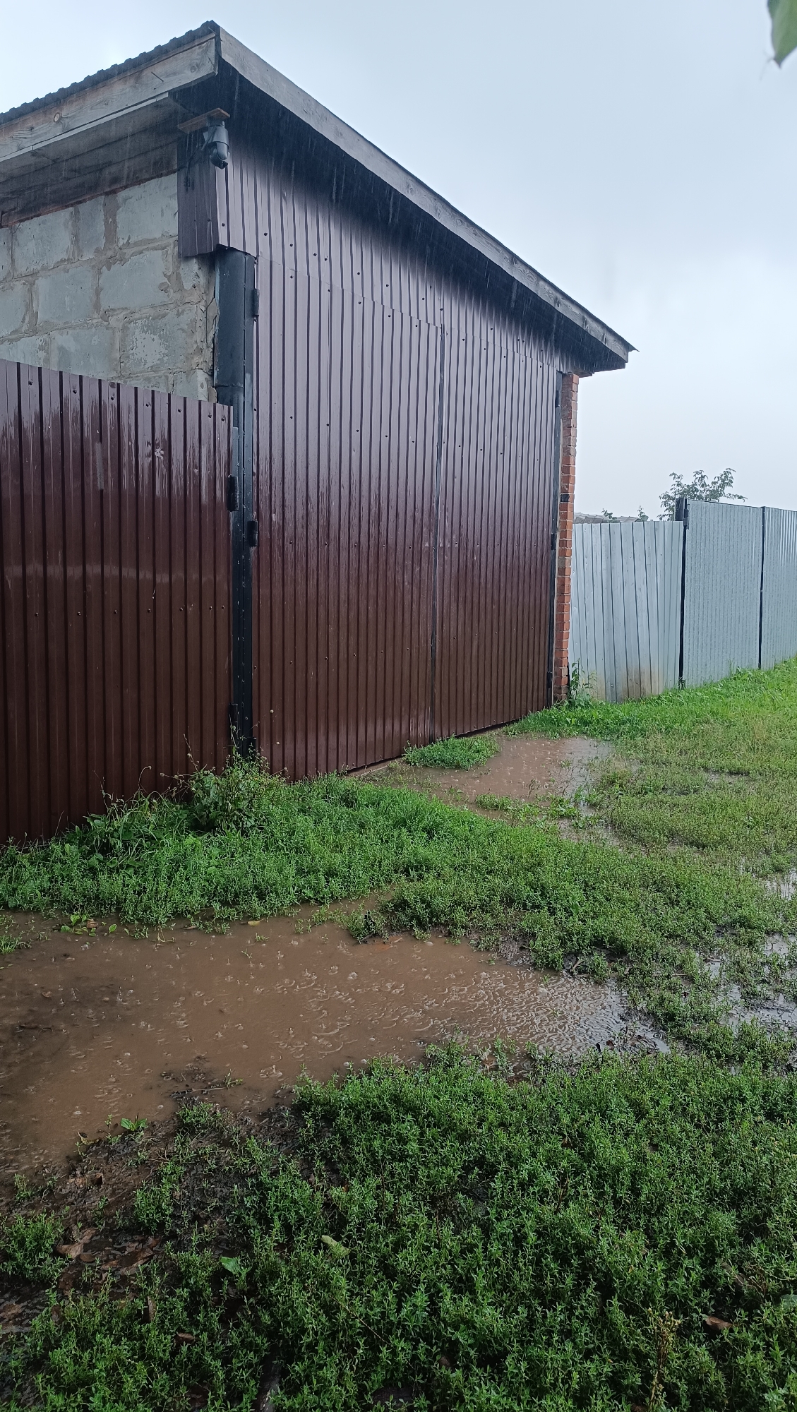 The image depicts an overcast, rainy day in a rural or suburban setting, likely in Karsha, Russia. A corrugated metal building, painted a dark brown, dominates the left side of the frame, its roofline angled upwards towards the right. The building's exterior wall is constructed of cinder blocks in its upper portion, with a dark downspout extending vertically along the corner. In front of the building is a brown metal fence. To the right of this, a lighter, silver corrugated metal fence runs parallel. The ground in the foreground is covered with green grass and shows several large puddles of muddy water, indicating recent or ongoing rainfall. The sky is a uniform, dull gray, typical of rainy weather. No people are visible in the image. The scene is focused on the exterior of the building, the fences, and the wet, muddy ground.
