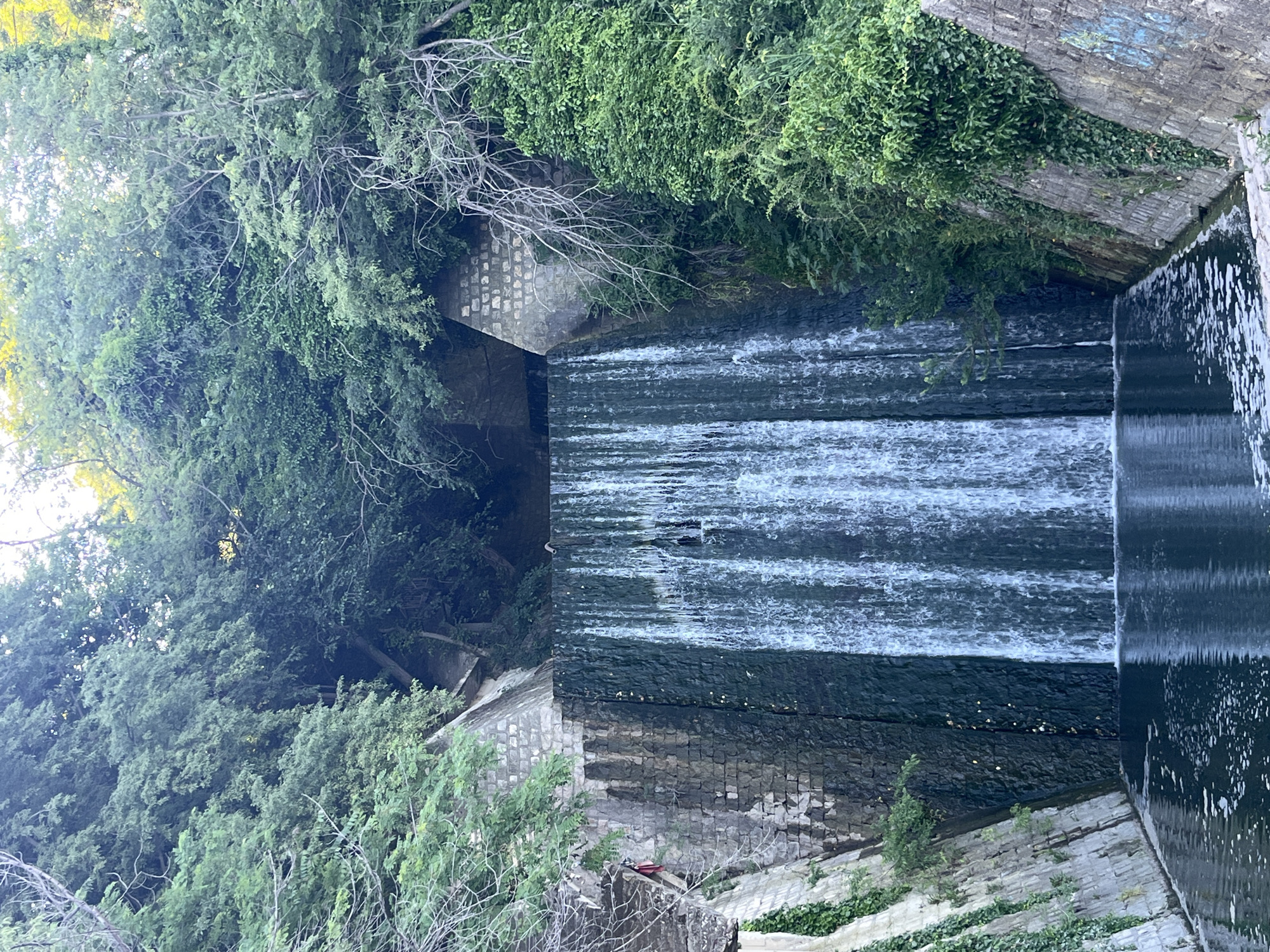 The image shows a man-made waterfall cascading down a stone structure, surrounded by lush green trees and vegetation. The water flows into a dark pool at the bottom. The stone structure appears to be part of a canal or waterway, with walls lined with stone blocks and some areas covered in ivy. The overall setting suggests a park or a landscaped area within a city. The scene is likely located in Varna, Bulgaria, known for its numerous green spaces and natural beauty.