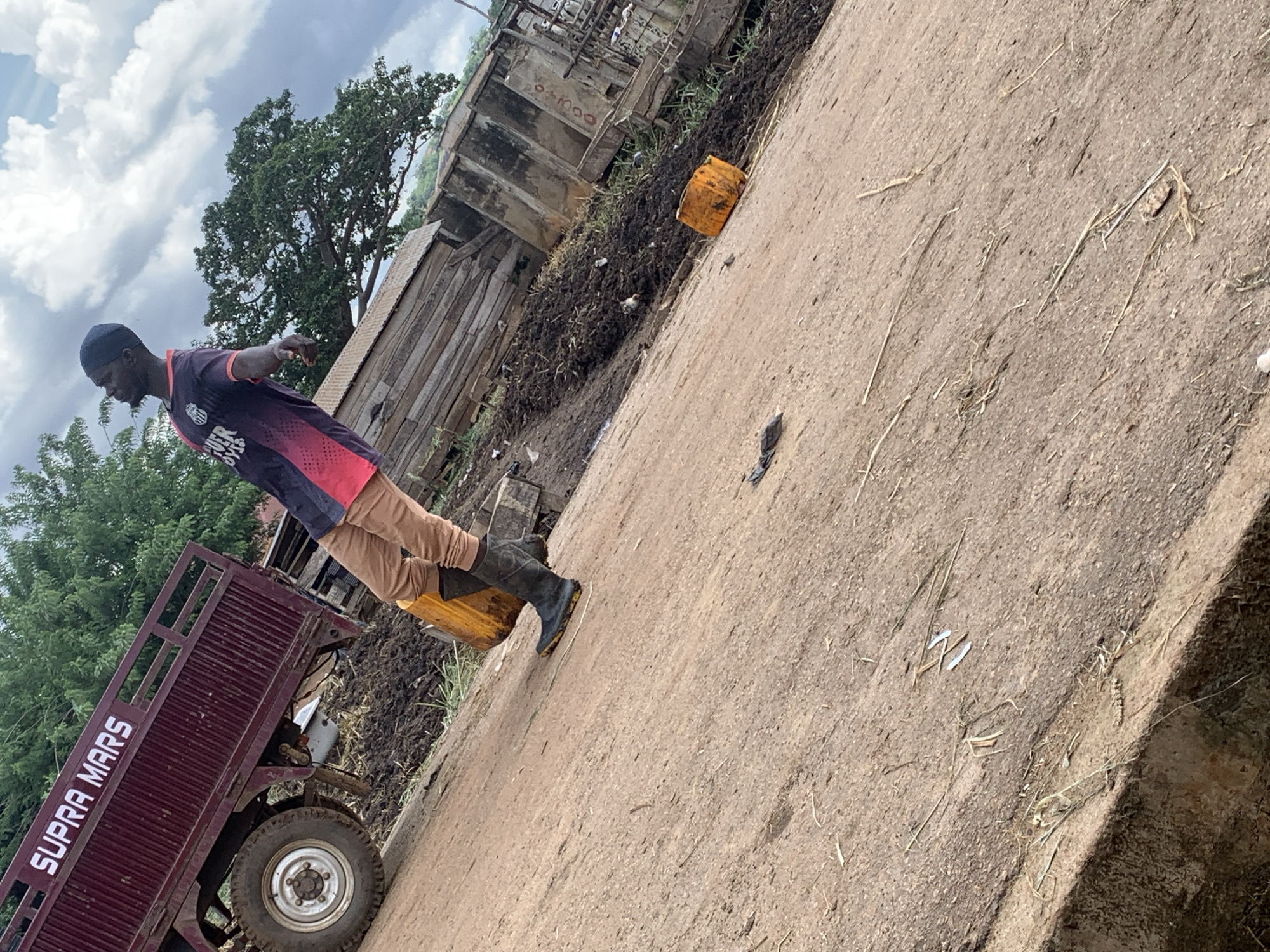 This media file depicts a scene in Akure, Nigeria, under a cloudy sky. The main subject is a man wearing a dark beanie, a multi-colored athletic shirt with pink and navy accents, tan trousers, and dark boots. He is positioned on the left side of the frame, leaning slightly back with his arms outstretched.
In the foreground, a maroon flatbed truck with the text "SUPRA MARS" in white block letters is visible on its side. The truck has a large tire on the right side of the frame.
The ground appears to be a dirt or gravel path, scattered with dry grass and other debris. To the right of the man, a yellow container is placed on the ground. In the background, there are structures that look like makeshift buildings or stalls, constructed with wood and corrugated metal. A pile of dark, possibly organic material, is also present near these structures, with some grass growing through it.
The overall impression is of a rural or semi-urban environment, possibly a market or work area, given the presence of the truck and the surrounding structures. The lighting suggests it is daytime, but the clouds indicate overcast weather. The man's posture could suggest he is either posing or involved in an activity related to the truck or the surrounding environment. There are no overt emotional cues visible.