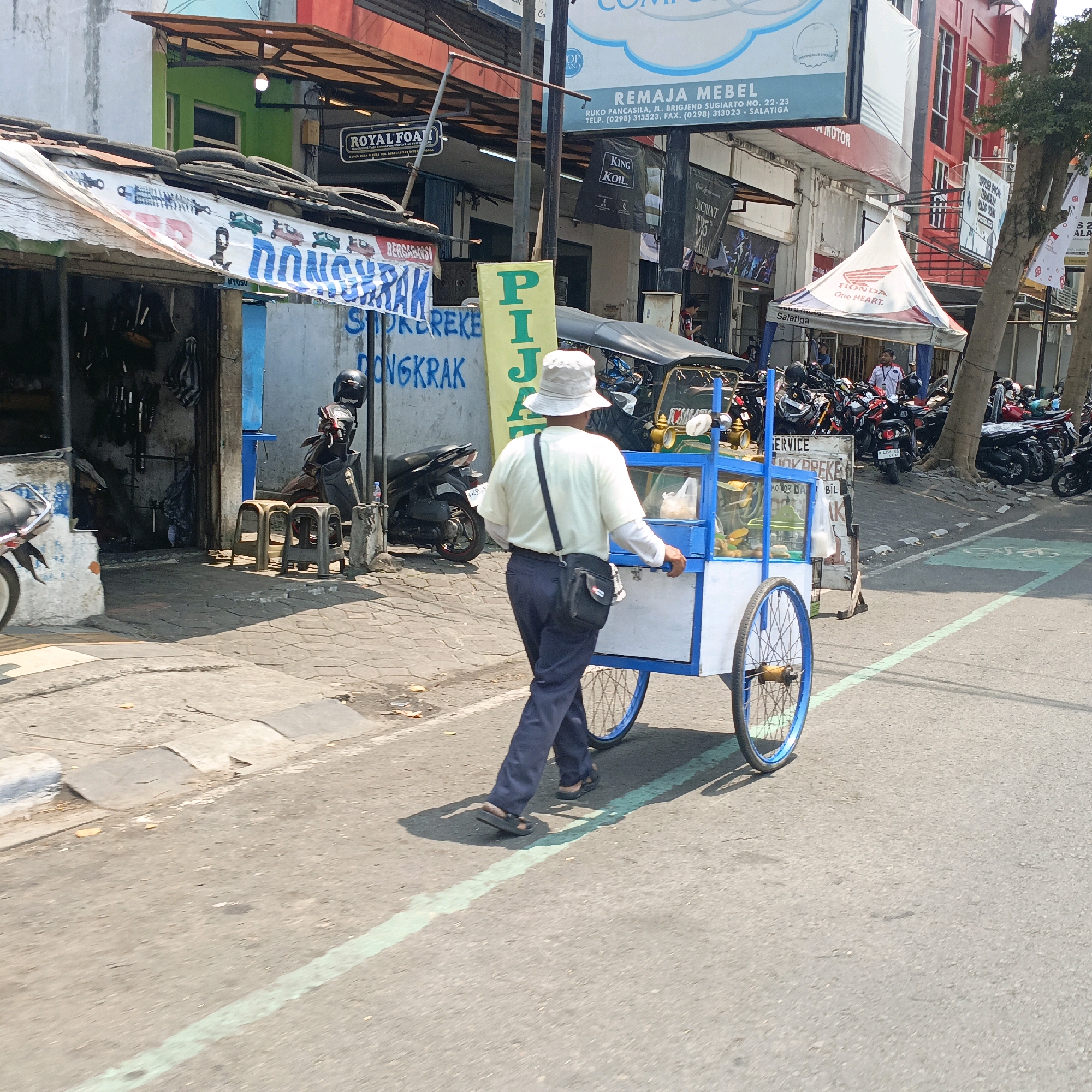 The image depicts a street scene in Salatiga, Indonesia, characterized by its bustling urban environment. In the foreground, a man wearing a light-colored bucket hat, a long-sleeved cream shirt, and dark blue pants pushes a mobile food cart. The cart is blue and white, with glass enclosures displaying food items, and has two large spoked wheels. He is walking on a paved street marked with a green line, possibly indicating a bike lane or pedestrian crossing. Behind him, a motorcycle repair shop is visible, with a banner displaying text and graphics, including images of shock absorbers and motorcycles. The shop's exterior is weathered, with a corrugated metal roof covered in what appear to be old tires. Several motorcycles are parked in front of the shop, and two plastic stools are placed on the sidewalk. Further down the street, a Honda dealership tent is set up, with numerous motorcycles parked underneath and in front of it. Various signs and banners are present on the buildings lining the street, advertising businesses such as "REMAJA MEBEL" and "ROYAL FOAM." The overall impression is of a lively commercial area on a sunny day.
