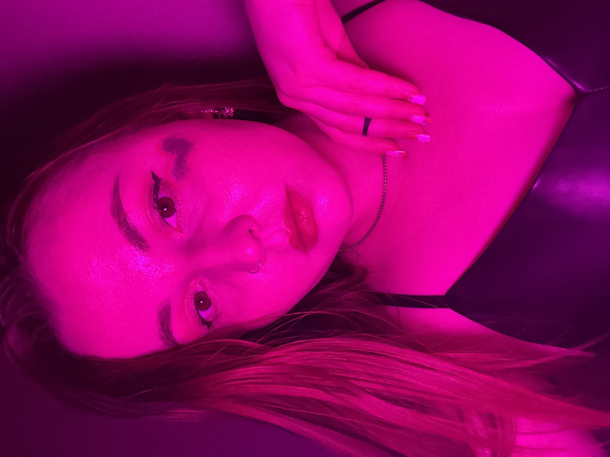 A female is shown in a close-up shot, lying down. Her face is directed toward the camera with her head tilted. The lighting is predominantly magenta. She wears makeup including lipstick, eyeliner and a design above her eyebrow. She also has a nose piercing, jewelry in her hair and is wearing a thin chain around her neck. Her hand is resting on her neck with a ring on her finger. A black garment covers part of her body and she has red/pinkish hair spread out behind her head. The background is indistinct. There is no information in the image to suggest a location of Izhevsk, Russia.
