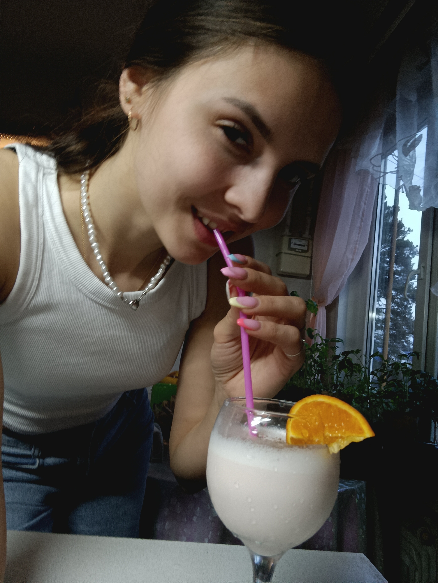 A young woman is depicted in a close-up, indoor shot, looking towards the viewer with a friendly smile. She is captured from the chest up, leaning slightly forward, with her dark hair pulled back. She wears a white ribbed tank top and a pearl necklace adorned with a small, heart-shaped pendant. A gold hoop earring is visible on her right ear, and a silver ring is on her left ring finger. Her long, manicured nails feature a colorful design with pink bases and multi-colored tips in shades of yellow, blue, and orange.
The woman is actively drinking from a bright pink straw, which is immersed in a stemmed glass filled with a light, frothy beverage, likely a milkshake or smoothie. The drink is garnished with a vibrant orange slice.
The background shows a window draped with light-colored, sheer curtains, possibly with a pink tint. Beyond the window, the bare branches of a large tree are visible, suggesting a cold season. Below the window, several small potted plants are arranged. To the left of the window, a white utility meter or box is mounted on the wall. Part of a dark surface or table is in the immediate foreground, and a patterned tablecloth is discernible further back. The lighting indicates it is daytime, with natural light illuminating the scene. The woman's expression conveys a sense of cheerfulness and contentment. There is no visible text in the image.