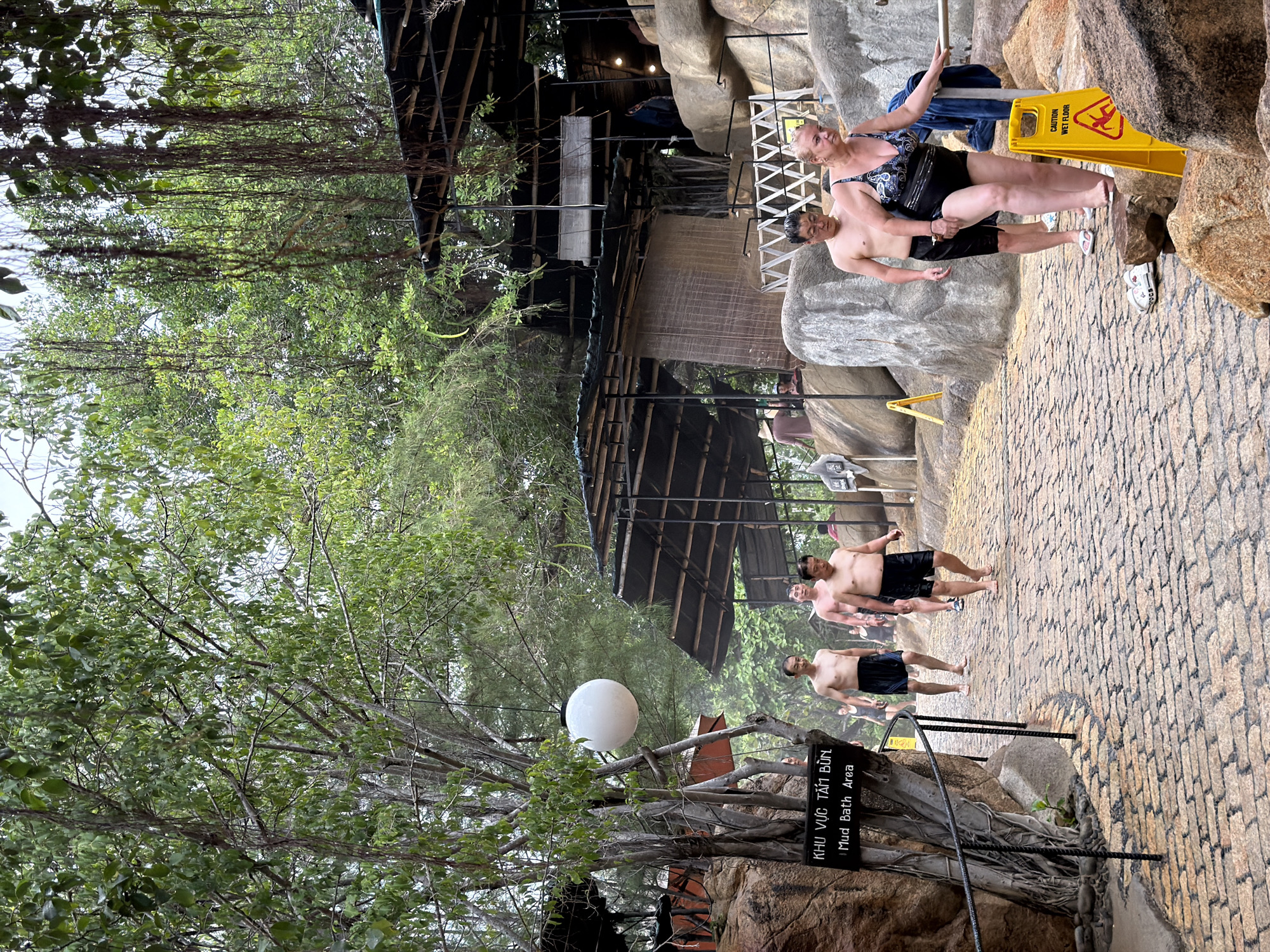 The image depicts an outdoor pathway at a mud bath facility in Nha Trang, Vietnam. The pathway is paved with rough, reddish-brown bricks and is slightly wet, suggesting recent rain or mist. Several people are walking along the path, most of them appearing to be tourists. In the foreground, a woman in a black and blue patterned swimsuit is walking towards the right, holding onto a railing. Behind her, a man with glasses, wearing black swim shorts, walks in the same direction. Further up the path, several men in swim shorts are walking away from the camera.
The environment is lush with vegetation, featuring large trees with hanging vines and dense green foliage. A large, white spherical lantern hangs from a tree branch on the left side of the path. To the left, a dark wooden sign with white text reads "KHU VUC TAM BUN" and "Mud Bath Area." In the background, there are covered structures with open sides, likely changing rooms or rest areas, built with dark roofing and wooden supports, nestled amongst large, grey rocks. A bright yellow "CAUTION WET FLOOR" sign stands on the right side of the path. The overall atmosphere suggests a humid, tropical setting within a recreational park.