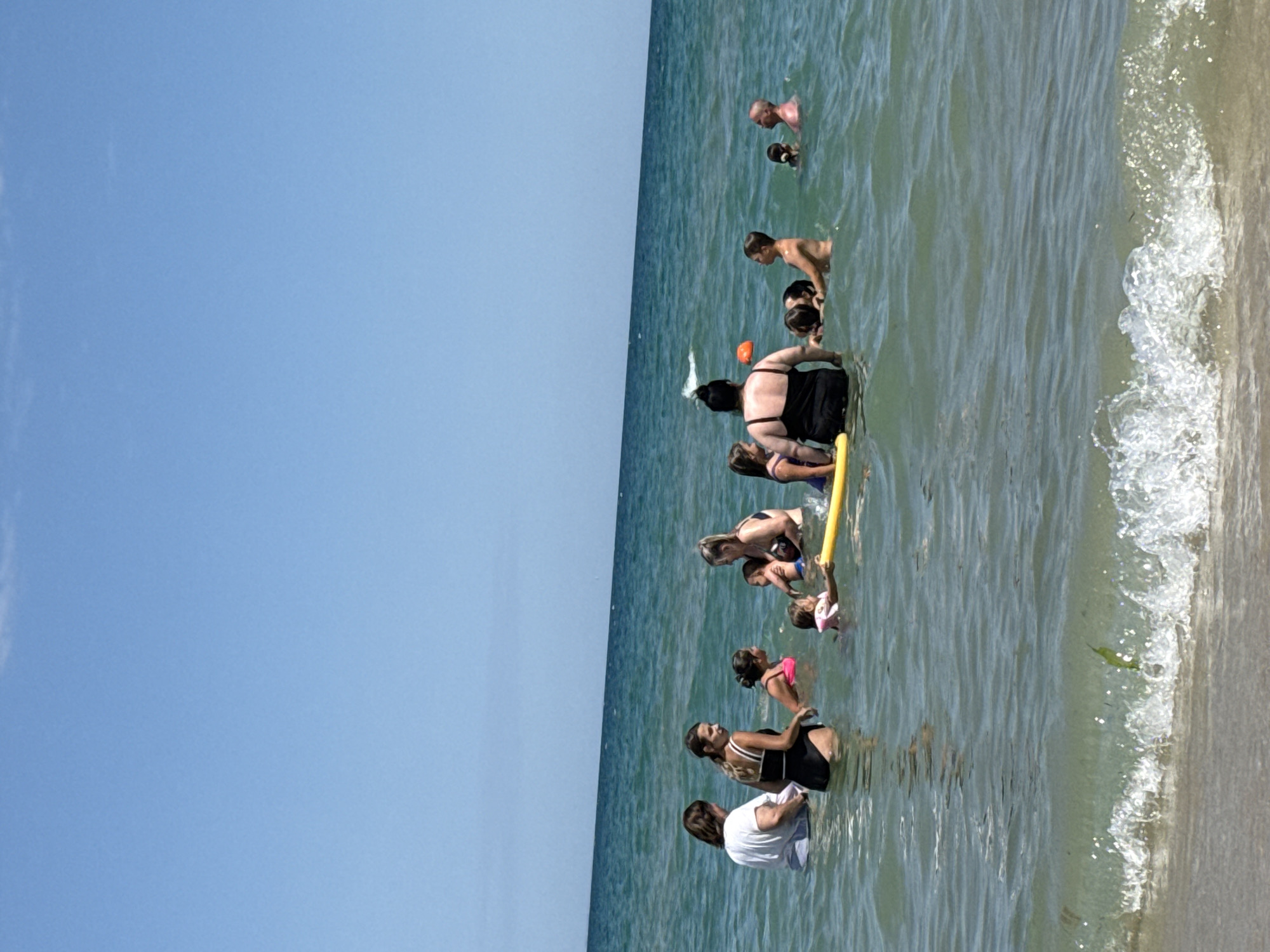 This is a photograph taken on a sunny day at the beach in Kranevo, Bulgaria. A group of people, including adults and children, are enjoying the water. Several individuals are in the sea, with some wading in the shallows and others further out. There is a yellow floatation device in the water, and a small orange object can be seen in the distance. The sea is a clear blue-green, with small waves breaking near the shore. The sky is bright blue with a few wispy clouds. The sand of the beach is visible in the foreground.