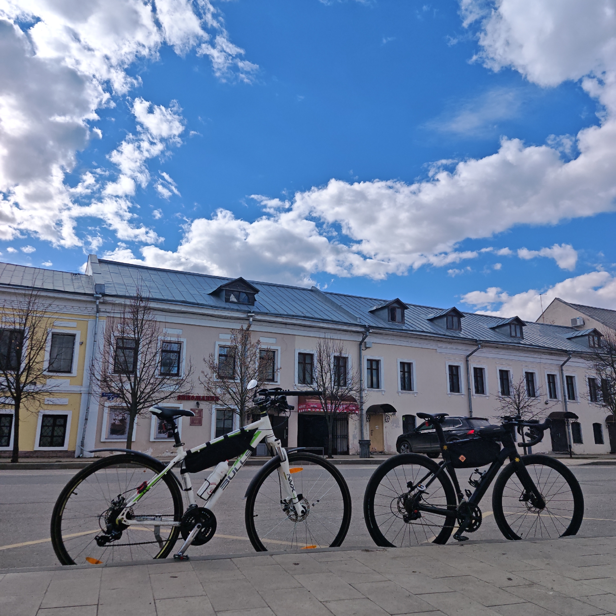 Two bicycles are parked side-by-side on a curb in Rostov, Russia. The bicycle on the left is white with green accents and has a black storage bag attached to the frame. The bicycle on the right is black and also has a black storage bag attached to the frame. Behind the bicycles is a row of two-story buildings with a light-colored facade and a metal roof. The trees are bare. The sky is blue with scattered white clouds. A black car is parked behind the black bicycle. The ground appears to be paved.