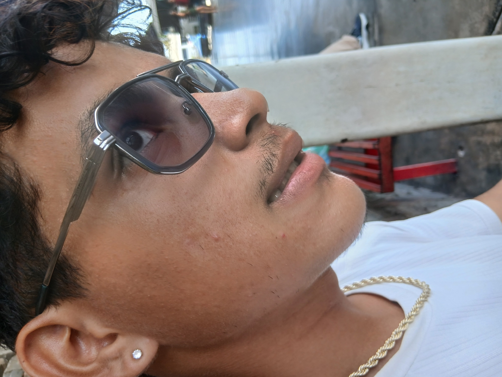 A close-up shot of a man wearing sunglasses and a gold chain, lying down. He has dark hair and a small earring. The background is blurred, but possibly shows a street scene. Without more specific identifying features in the background, determining the location in Juan Griego, Venezuela, is not possible.