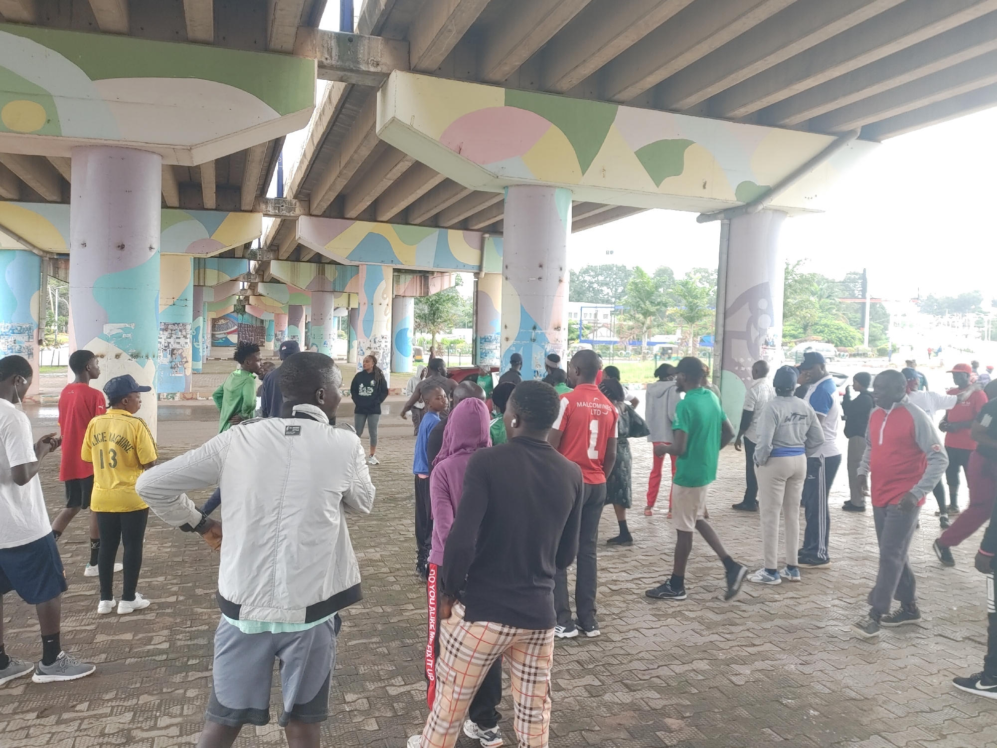 The image depicts an outdoor gathering of approximately twenty people standing under a concrete overpass with brightly painted pillars. The setting is likely an urban area, possibly in Jos, Nigeria, given the presence of a billboard in the background featuring text in what appears to be Hausa language, and typical Nigerian architectural elements.
The individuals are of various ages and genders, dressed in casual attire, with many wearing athletic or casual wear. Some are standing in groups, while others are more spread out. The ground is paved with interlocking bricks.
The overpass structure dominates the upper portion of the image. The pillars supporting it are decorated with colorful, abstract murals featuring geometric shapes in pastel colors like green, purple, yellow, and pink. This suggests a deliberate effort to beautify or make the underpass area more visually appealing.
In the background, beyond the overpass, trees and some buildings are visible, indicating it's not an isolated area. There are also some poles and what might be streetlights or infrastructure elements. The overall atmosphere appears to be informal and relaxed, with people congregating in what could be a public space.