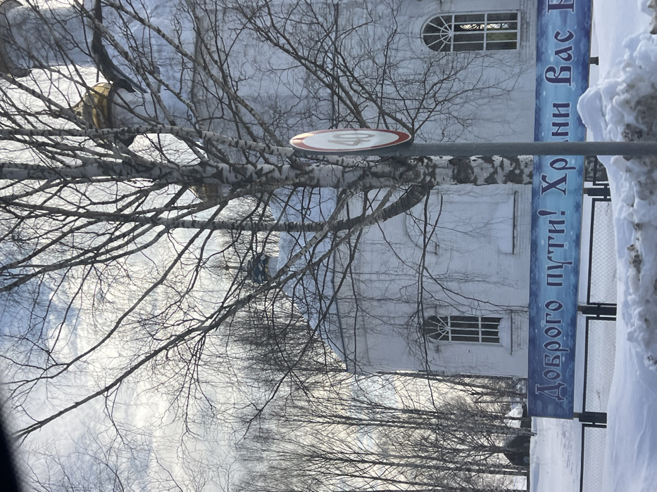 A winter scene in Полозово, Russia, captured under a partially cloudy sky. The foreground shows an accumulation of snow, forming uneven drifts. A metal fence is partially visible behind the snow, supporting a blue banner with white Cyrillic text that reads, "Доброго пути! Храни Вас" (Have a good journey! May God keep you). Dominating the midground is a white-painted church or temple with a prominent golden onion dome and a smaller blue dome, both topped with crosses. The church features arched windows with vertical bars. Its roof and ledges are covered with snow. A bare-branched birch tree stands centrally, partially obscuring the church's facade. To the right of the birch tree, a circular speed limit sign, white with a red border and displaying "40" in black, is mounted on a grey pole. The background consists of numerous bare deciduous trees, suggesting a wooded area. No individuals or active subjects are present.