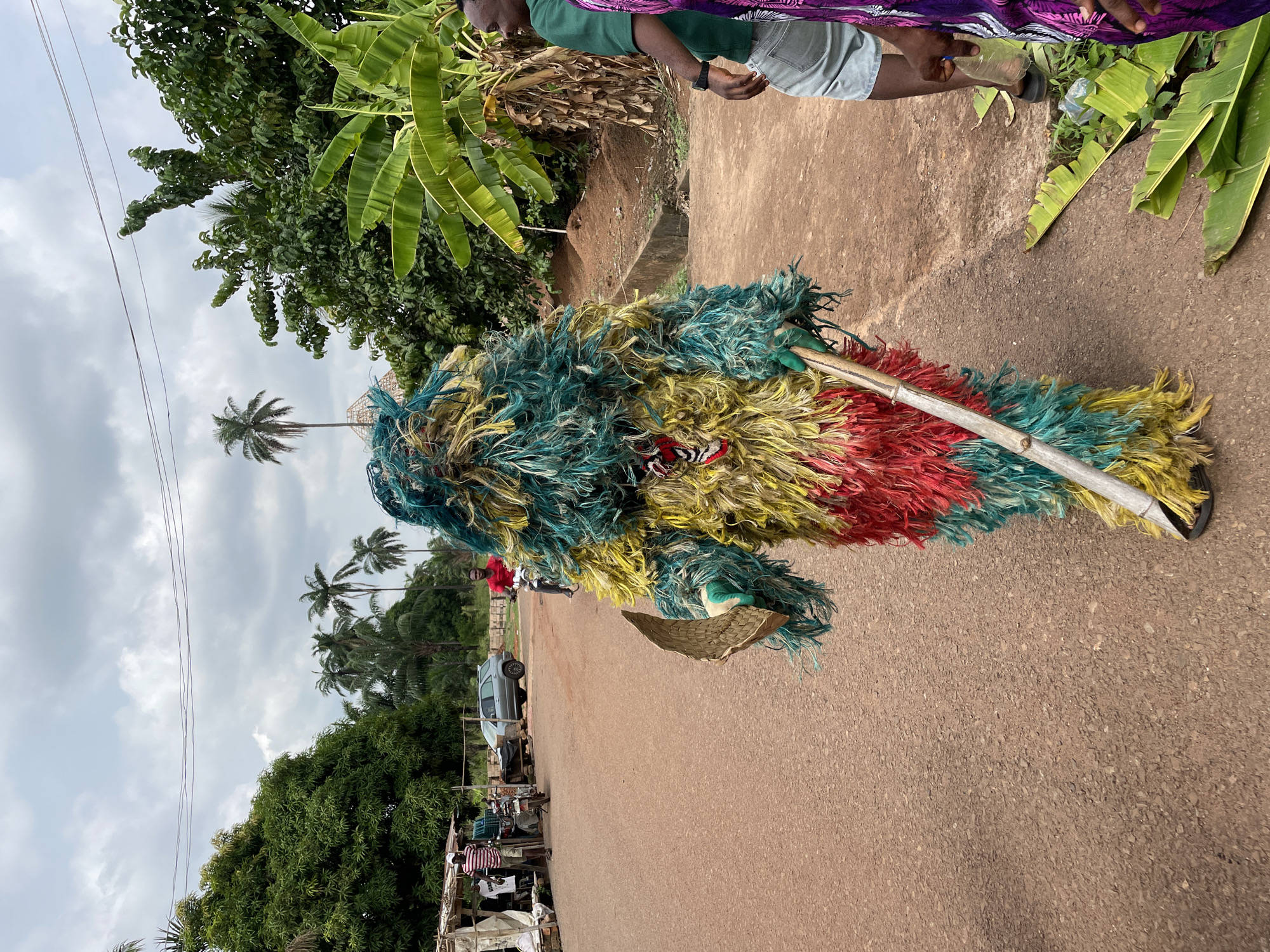 A masquerade figure, predominantly composed of vibrant blue, yellow, and red synthetic fibers, is seen on an asphalt road in Agba Umana, Nigeria. The masquerade holds a small, woven basket and a long, smooth pole. An individual in a green shirt and denim shorts stands at the edge of the road next to banana trees. Behind the masquerade, a gray sedan is parked near market stalls. Palm trees rise in the background under a cloudy sky.