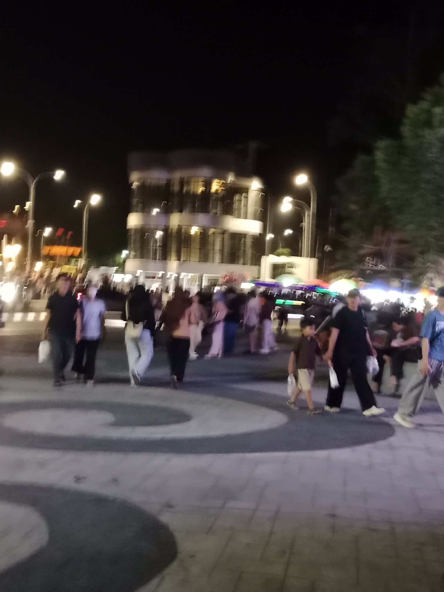The image depicts a street scene at night in Palangka Raya, Indonesia. A group of people are walking on a paved area with circular patterns. Several streetlights illuminate the scene, and a multi-story building with lit windows is visible in the background, as are trees. Some individuals are carrying bags. The lighting suggests it is nighttime.