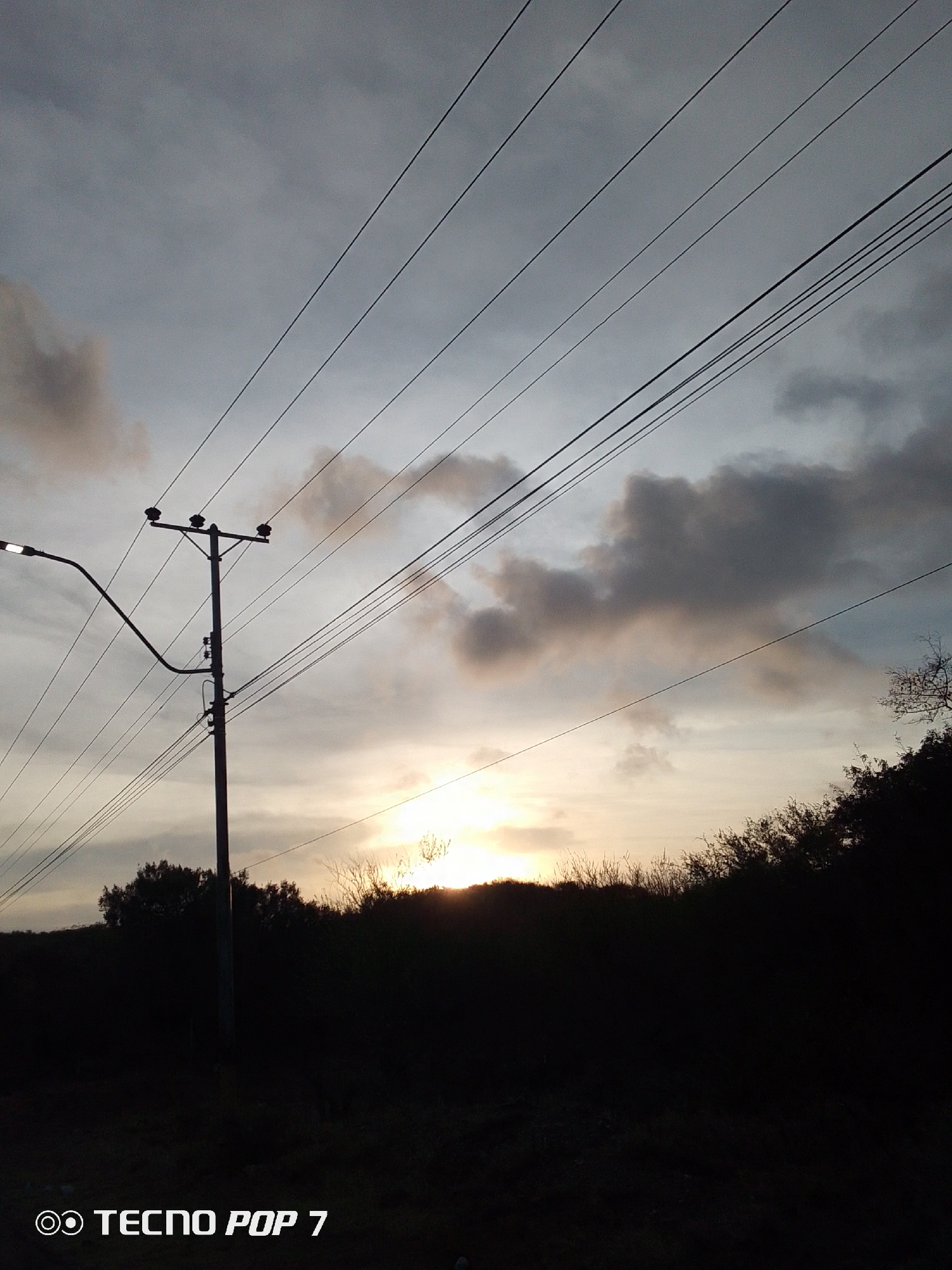 The image depicts an outdoor scene, likely taken at dusk or dawn, characterized by a low-angle view towards the horizon. In the foreground, there's a dark, silhouetted landscape with dense vegetation obscuring most details. A utility pole stands prominently to the left of the center, supporting power lines that extend diagonally across the frame towards the upper right corner. A streetlight is attached to the pole, although it's unclear if it is illuminated. Above the silhouetted landscape, a band of bright light suggests the presence of the sun, either setting or rising. The sky is overcast with grey and white clouds. The location is potentially in Altagracia, Venezuela, though this cannot be forensically verified from the image alone. The overall impression is one of a rural or suburban setting. The image also has the text "TECN POP 7" in the lower-left corner.
