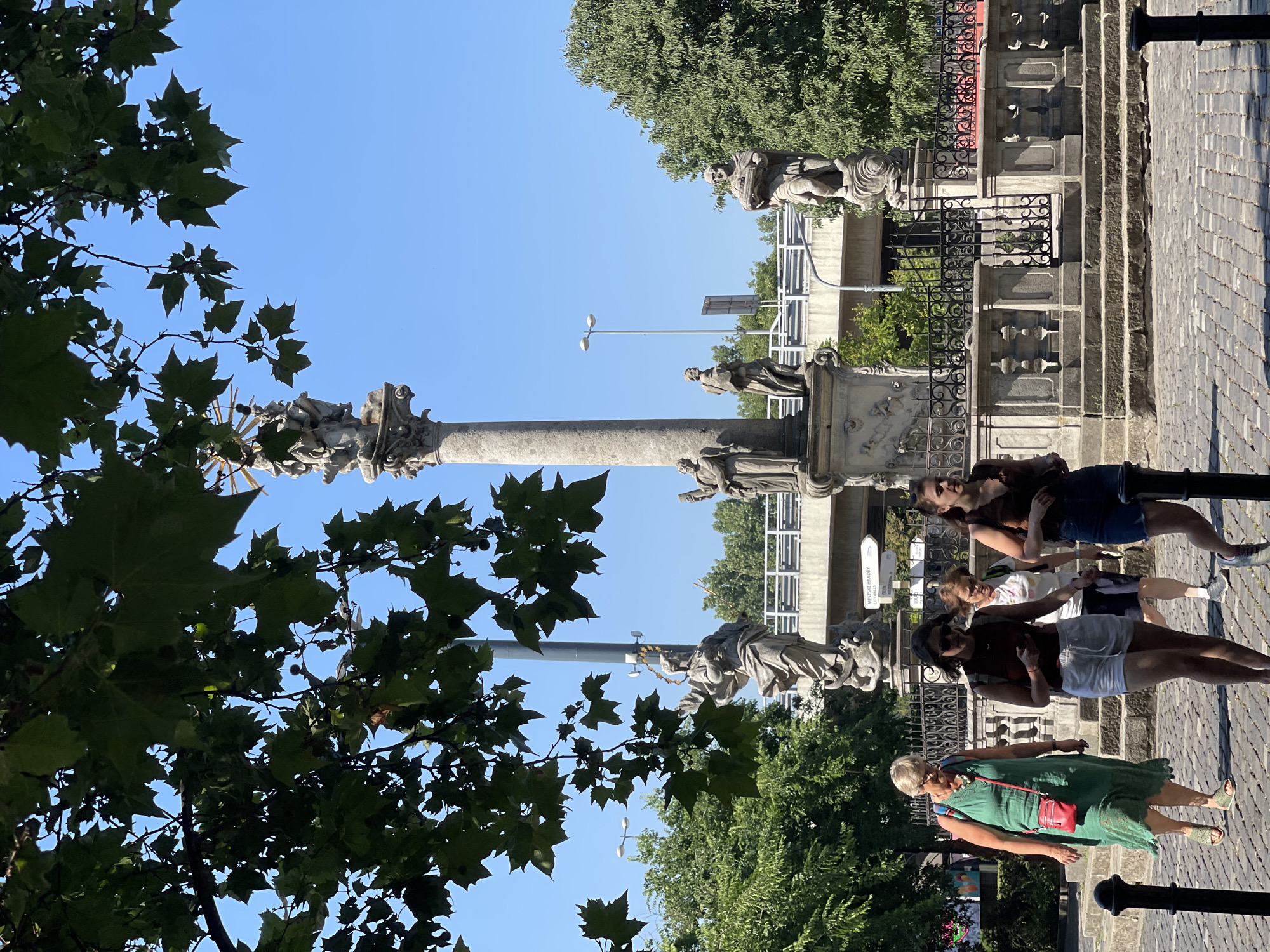 In Bratislava, Slovakia, on a bright, sunny day, a group of women walk along a cobblestone street in front of a historical monument. The monument, likely the Plague Column or a similar Baroque structure, features several stone statues and is topped by a further sculptural element. Lush green trees frame the top of the image, partially obscuring the clear blue sky.
The women, four in total, are captured mid-stride. The woman on the far left wears a flowing green dress and a red crossbody bag, while the woman next to her sports white shorts and a dark top, looking down at her phone. Behind them, another woman in a white t-shirt and dark shorts is visible, and the fourth woman, in a dark sleeveless top and denim shorts, is in the foreground on the right. The background reveals a multi-level urban environment with a bridge structure and streetlights. The paved ground is characteristic of an older European city center.