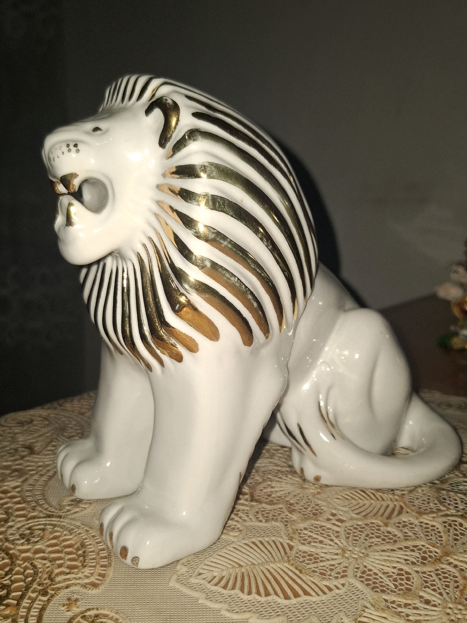 A white porcelain figurine of a seated lion with gold accents is positioned on a lace-patterned doily. The lion has its mouth open, revealing teeth, and a mane detailed with gold stripes. The background is dark and out of focus, with a blurred hint of other objects on the right. The scene is indoors, but no specific location in Almaty, Kazakhstan can be identified from the image.
