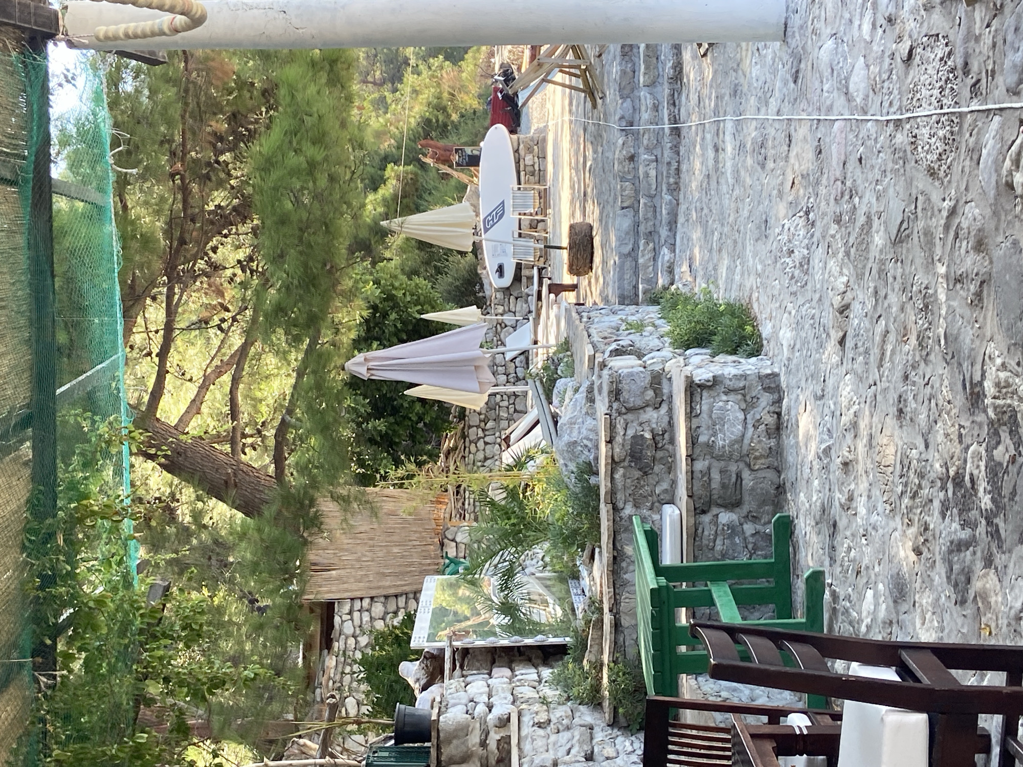 The image depicts an outdoor seating area near a body of water, possibly a beach or a harbor, in Prijevor, Montenegro. In the foreground, a dark wooden chair with a white cushion is visible. Behind it, a bright green wooden table and benches are situated on a stone-paved area.
The middle ground features a stone-built tiered structure with various plants and a large framed artwork leaning against it. Several beige patio umbrellas are set up, and a white surfboard is placed upright on a stone pedestal, supported by two chairs.
The background is dominated by lush green trees and foliage, with a stone wall and more greenery further back. The overall atmosphere suggests a relaxed, seaside establishment. The lighting indicates it is daytime, with sunlight filtering through the trees, creating dappled shadows.