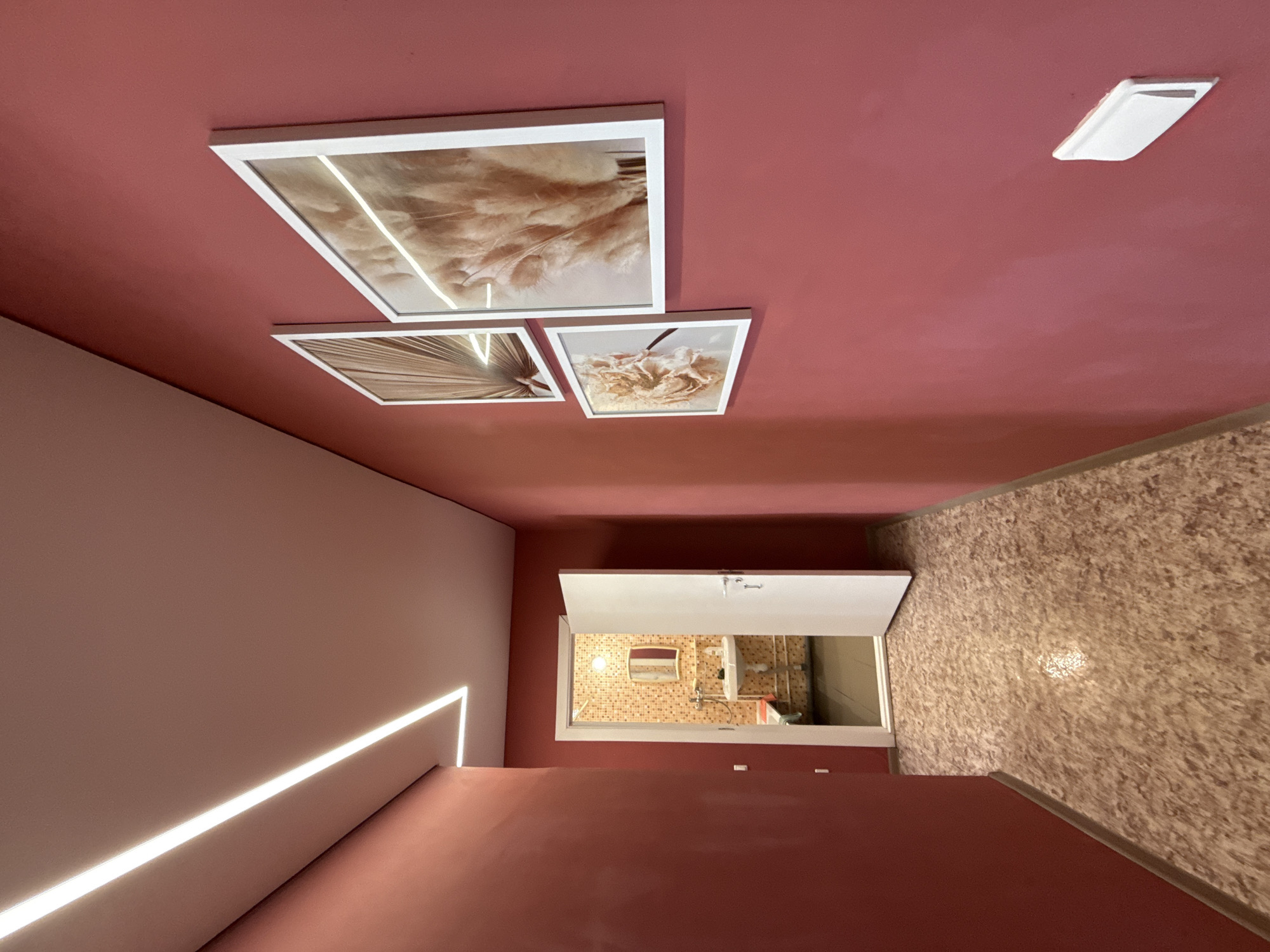 The image shows a hallway with red walls. There is a door open to a bathroom on the left. On the right wall, there are three framed pictures of dried flowers and plants. A linear light fixture is installed on the ceiling, creating an illuminated line that runs along the length of the hallway. The floor is covered with a speckled, light-brown linoleum. The overall scene is tidy and well-lit, suggesting an indoor residential or hospitality setting. The location is Valday, Russia.
