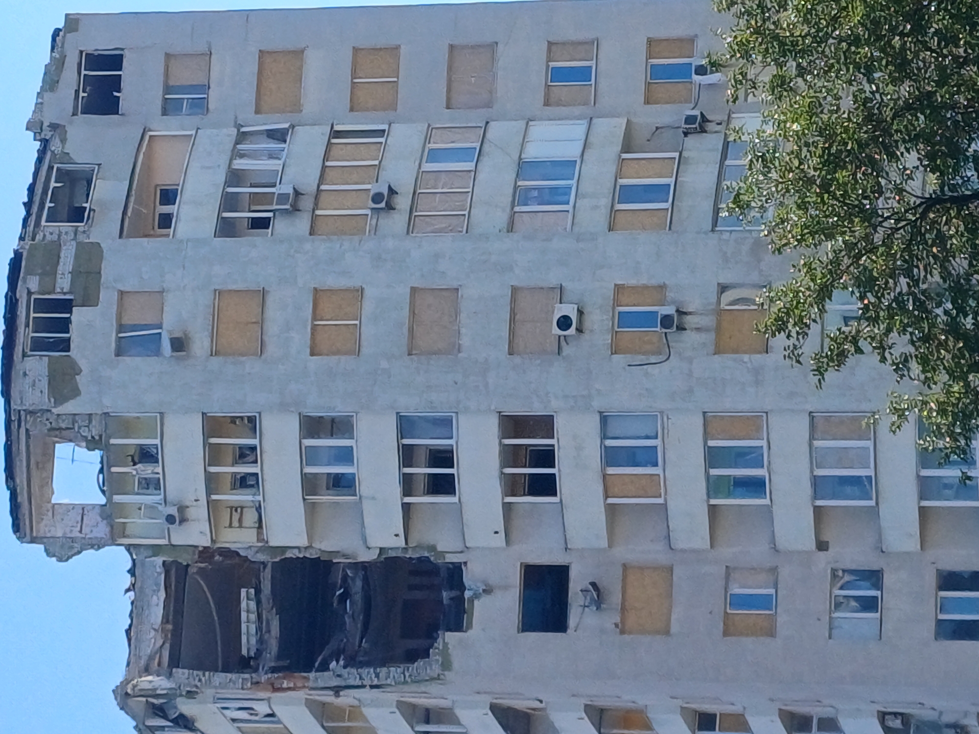 The image shows a residential building in Kyiv, Ukraine, with significant damage to its facade. The building appears to be several stories high, with a damaged section on the left side revealing the interior structure. Many windows are boarded up with wooden panels, while others are broken or missing. Several air conditioning units are visible on the exterior walls. Trees with green foliage are in the foreground, partially obscuring the lower part of the building. The sky above is clear and blue.