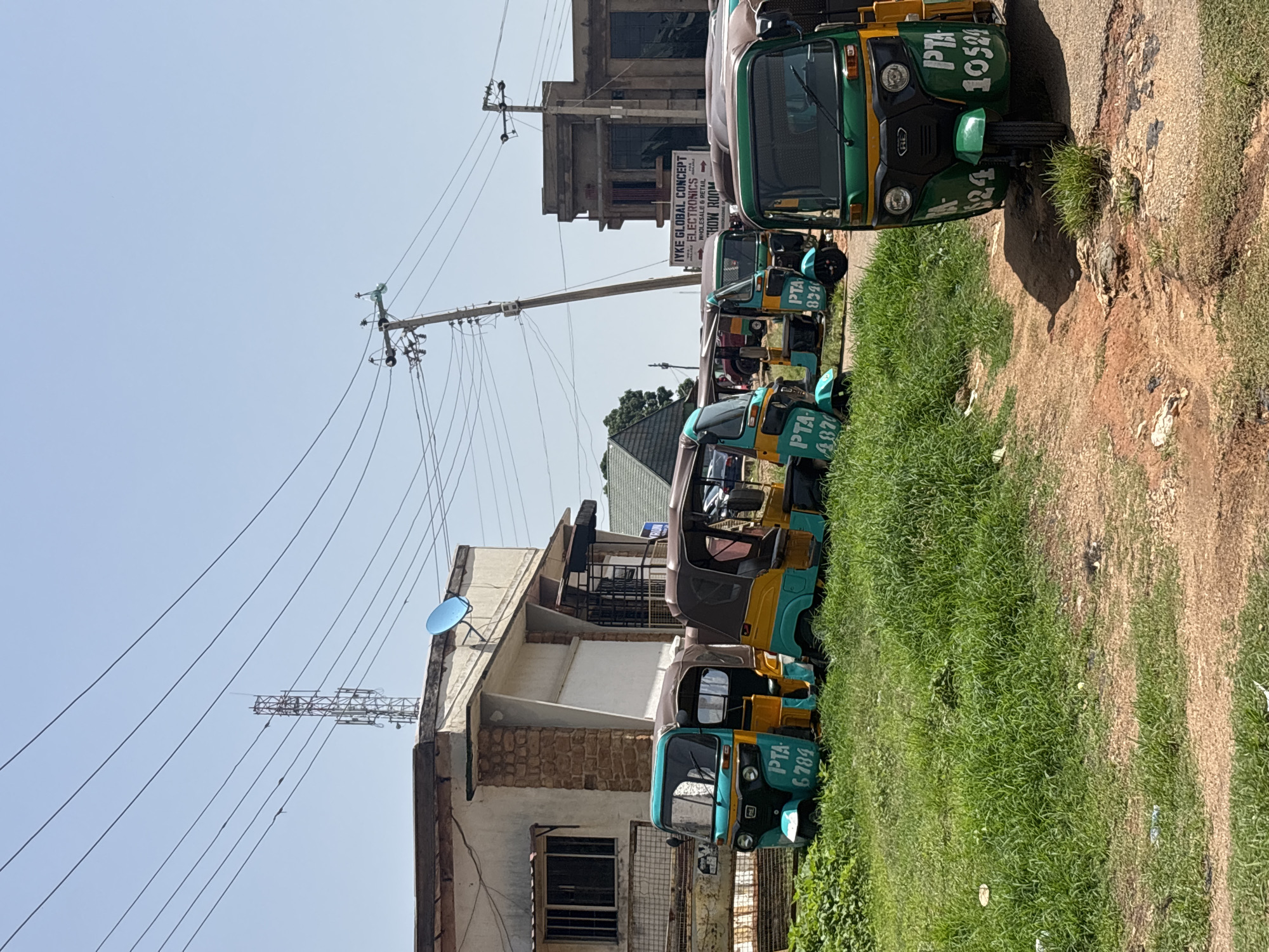 In Jos, Nigeria, on a sunny day, a row of colorful auto-rickshaws, known locally as Keke Napep, are parked on a dirt road next to a grassy area. These three-wheeled vehicles, primarily painted in shades of turquoise, yellow, and green, display license plates such as "PTA 6784," "PTA 4870," and "PTA 8340," and "PTA 1059." In the background, buildings line the street, including a light-colored structure with stone accents and a larger, darker building with signage indicating "JYKE GLOBAL CONCEPT ELECTRONICS WHOLESALE & RETAIL SHOW ROOM." Power lines and a telecommunications tower are visible against the clear blue sky. The scene suggests a typical urban setting in Jos, likely a commercial or transport hub.