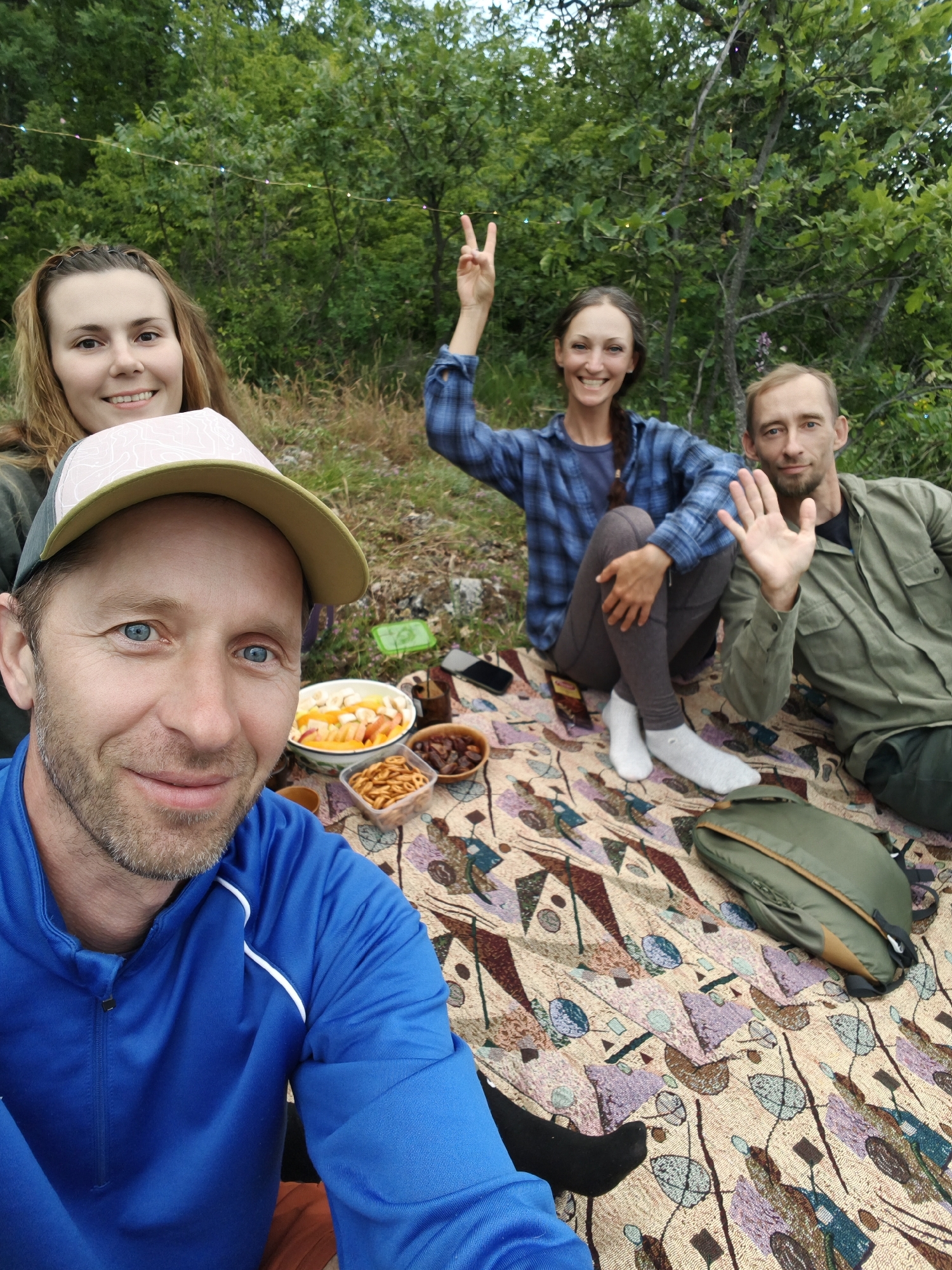 The image depicts an outdoor picnic scene with four individuals in Kiparisnoe, Ukraine.
In the immediate foreground, a man with light blue eyes, light brown facial hair, and a slight smile is taking a selfie. He wears a blue long-sleeved quarter-zip shirt with white piping on the shoulder and a two-toned baseball cap with a pale pink/white crown and olive green brim.
Behind him, to the left, a woman with long light brown hair is visible, smiling. To her right, a woman with long dark brown hair styled in a braid sits on a patterned blanket. She wears a blue and white plaid long-sleeved shirt and dark gray leggings, with white socks on her feet. She is holding her right hand up in a peace sign. To her right, a man with short light brown hair and faint facial hair sits on the blanket, wearing an olive green long-sleeved button-up shirt and dark green pants. He holds his left hand up in a waving gesture.
The group is seated on a large, patterned blanket featuring abstract geometric and organic shapes in shades of beige, brown, purple, blue, and green. Various food items are spread on the blanket: a white bowl of sliced fruit (including peaches and bananas), a clear plastic container of pretzels, and a small wooden bowl containing dates or dried fruit. A dark-colored mug and a small wooden implement are also visible. A black smartphone rests on the blanket near the woman in plaid, and a green and tan backpack or small duffel bag is positioned on the blanket in the lower right.
The background consists of dense green foliage, indicating a wooded or natural area. A string of multi-colored lights is strung horizontally across the trees. The ground beneath the blanket appears to be dirt and sparse grass.