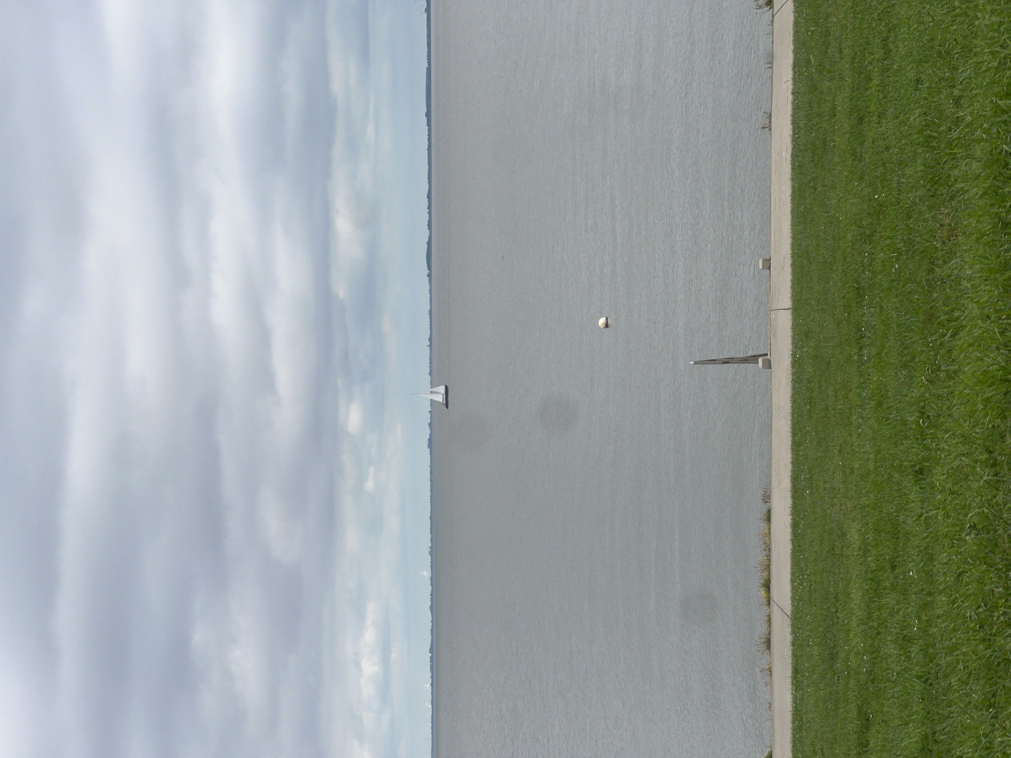 The image depicts a calm, overcast day at a body of water, likely a bay or sea, near Wilhelmshaven, Germany. In the distance, a solitary sailboat with white sails navigates the water. Along the horizon, wind turbines are visible, suggesting an industrial or coastal area. Closer to the foreground, a white buoy floats on the surface. A concrete embankment lines the water's edge, and beyond that, a grassy slope descends towards the viewer. The sky is filled with heavy, grey clouds. There are no people visible in the image.