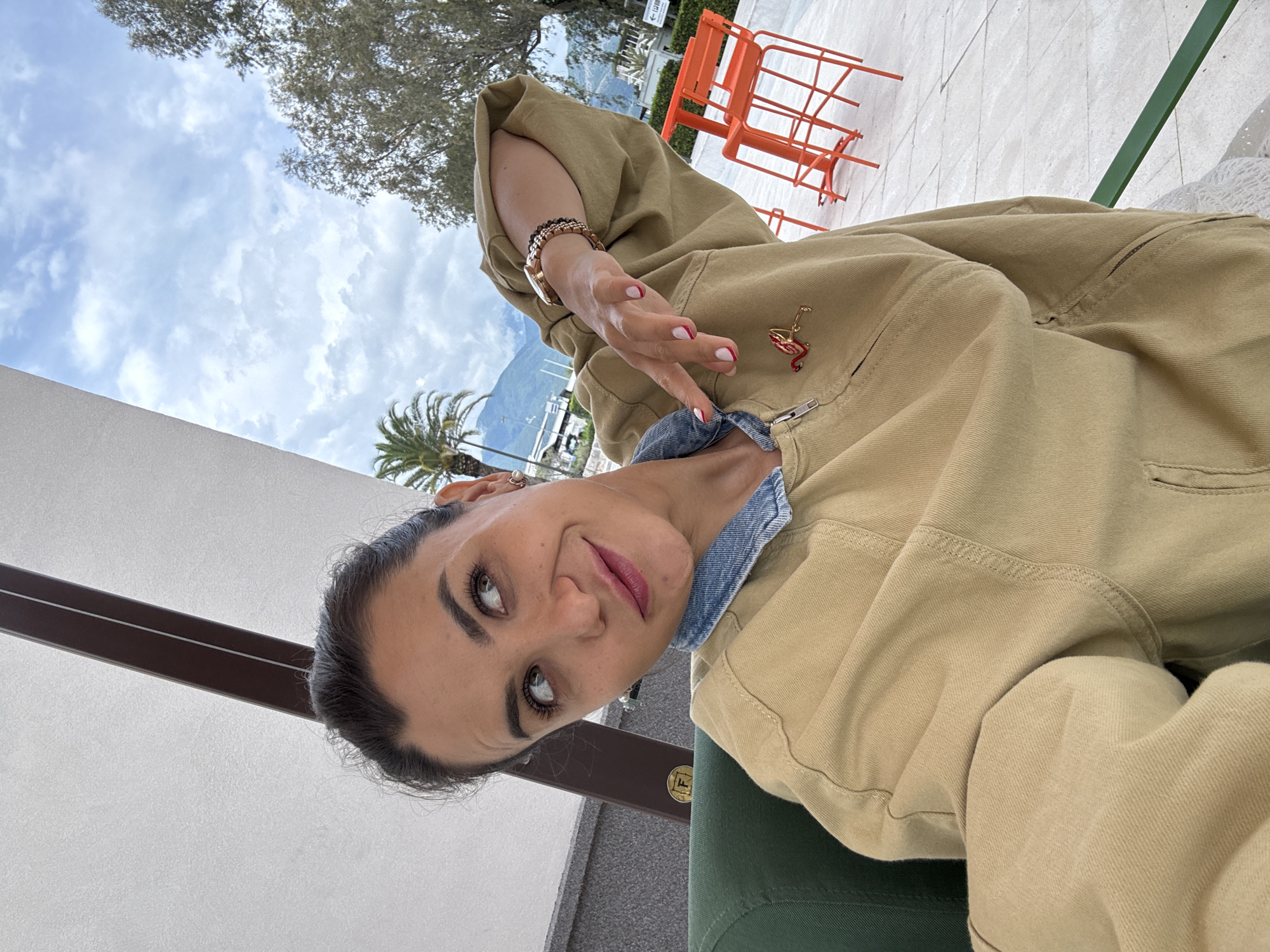 A woman is taking a selfie outdoors in Tivat, Montenegro. She's wearing a khaki jacket over a denim shirt. The woman has her hair pulled back, and she is wearing a gold watch and bracelets. Her fingernails are painted with a red and white design. Behind her is a backdrop of a blue sky with clouds, a palm tree, and mountains. There are orange chairs visible in the background. A white wall and part of a green chair are visible in the foreground.
