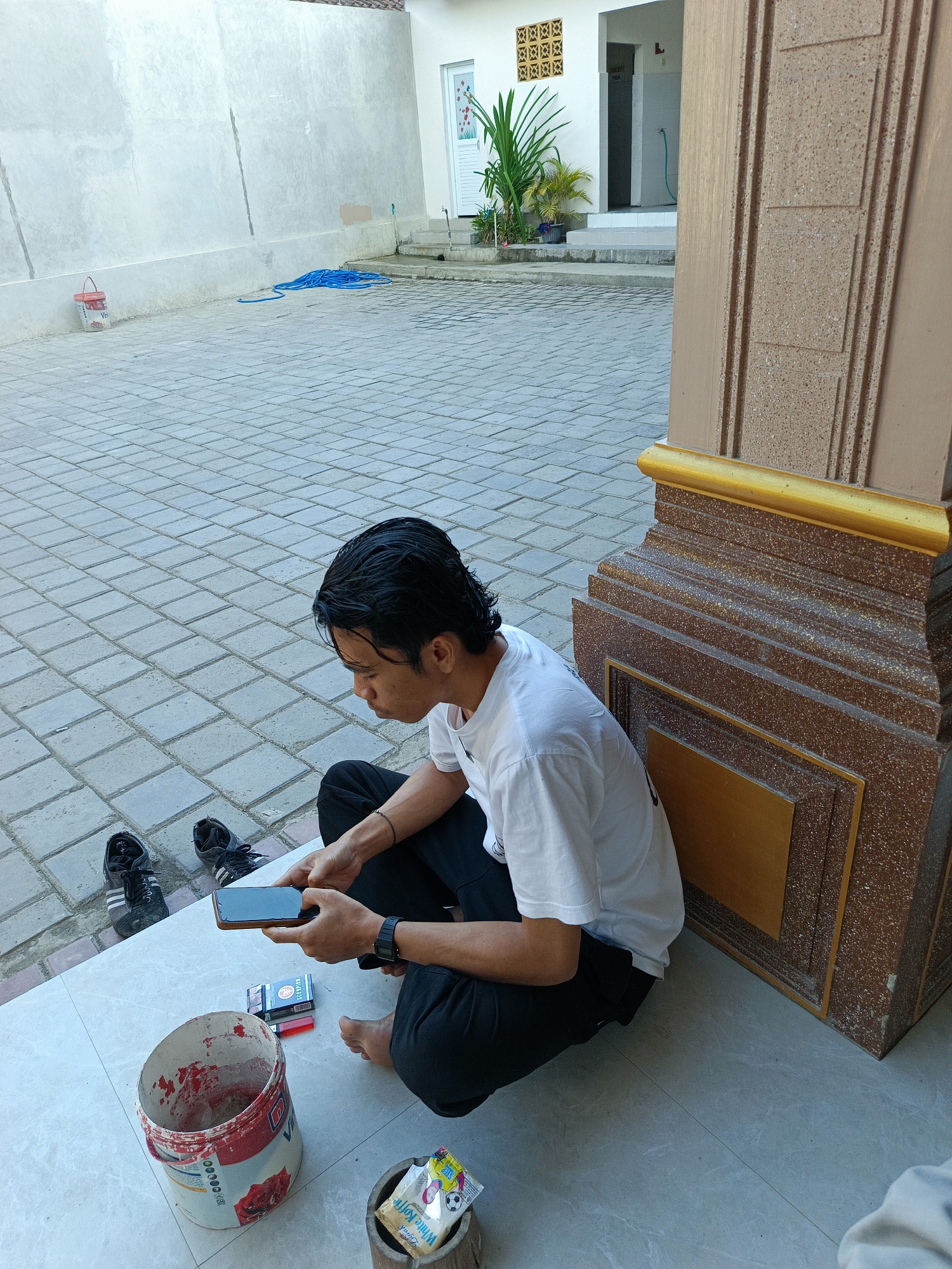 A young man wearing a white t-shirt and black pants is sitting on the ground, looking at his phone. He is crouched next to a large decorative pillar. In front of him are a pack of cigarettes, a lighter, and an open bucket with red paint inside. There are also a pair of black sneakers on the ground behind him. The man has dark hair and appears to be focused on his phone.
The setting is an outdoor courtyard paved with light gray tiles. A light-colored wall is visible in the background, with a doorway and a small garden. A blue coiled hose lies on the ground near the wall. The scene suggests a casual, everyday moment. The lighting indicates it is daytime, likely sunny given the shadows. The presence of paint suggests some kind of work or maintenance might be ongoing or has recently taken place.
The visible text on the cigarette pack reads "Gudang Garam". There is also text on the paint bucket, which appears to be a brand name in red and white. A packet of "White Koffie" is also visible near the paint bucket.