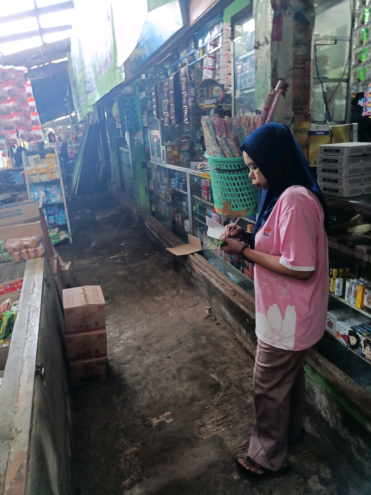 An overhead shot captures a street market scene in Mlawang, Indonesia. A woman wearing a blue hijab and a pink t-shirt with floral prints is standing on the right side of the frame, facing left. She is holding a pen in her right hand and appears to be writing on a piece of paper. In front of her, a green basket is filled with rolls of colorful paper.
The market stall is lined with shelves stocked with various items, including bottles and boxes. A narrow walkway separates the stall from a raised counter on the left, where boxes and packaged goods are displayed. The ground is a rough, gray surface, wet in places, indicating recent rain or cleaning. Sunlight streams in from the upper left, casting shadows and illuminating the dusty environment. The overall impression is that of a bustling, albeit somewhat cluttered, local market.