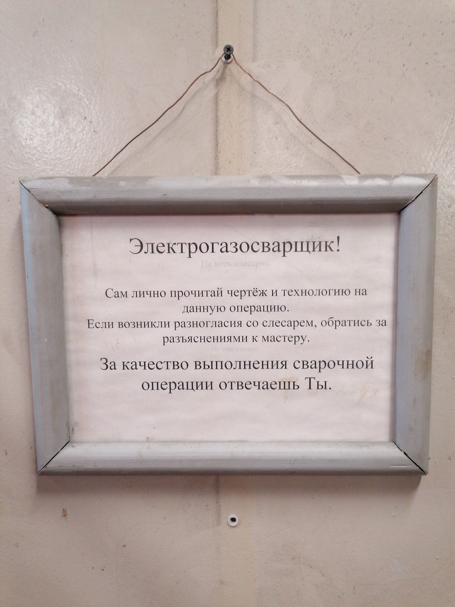 The image shows a framed sign hanging on a light-colored wall. The sign has a light gray frame and is suspended by a thin wire attached to the wall. The sign is written in Russian, likely a warning or instructions for an "electric gas welder" (Электрогазосварщик!). The sign instructs the welder to personally read the drawing and technology for the given operation, to address any discrepancies with a fitter to the foreman, and warns that the welder is responsible for the quality of the welding operation. The image suggests an industrial or workshop environment. The location in the city of Unknown, Unknown is indeterminate.