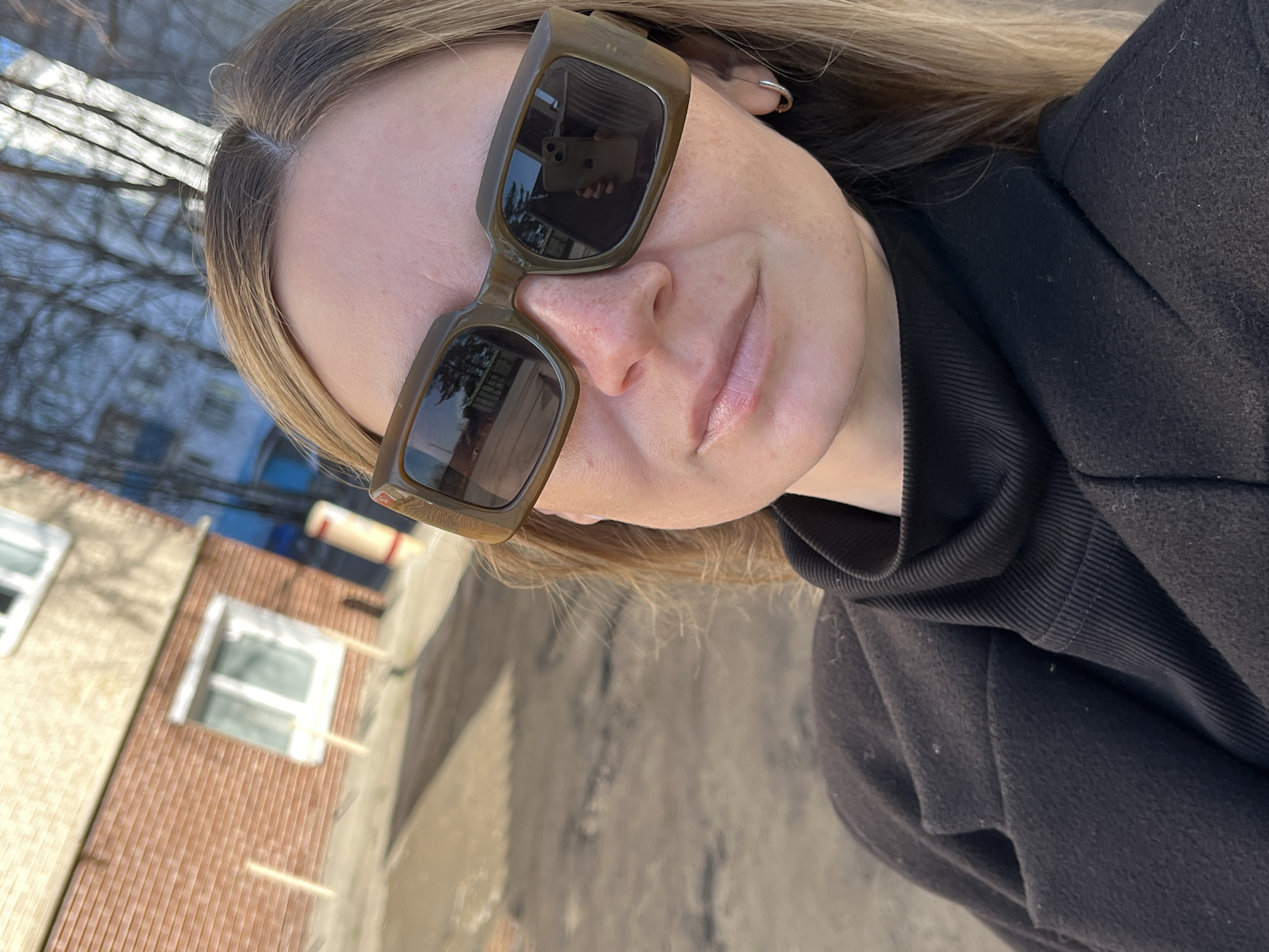 A woman is outdoors, wearing olive-green sunglasses, a black turtleneck, and a black jacket. She is in front of a brick building with white-framed windows, possibly in Chaikovsky, Russia. The sunglasses reflect the building and trees. The ground appears to be a concrete or asphalt surface.