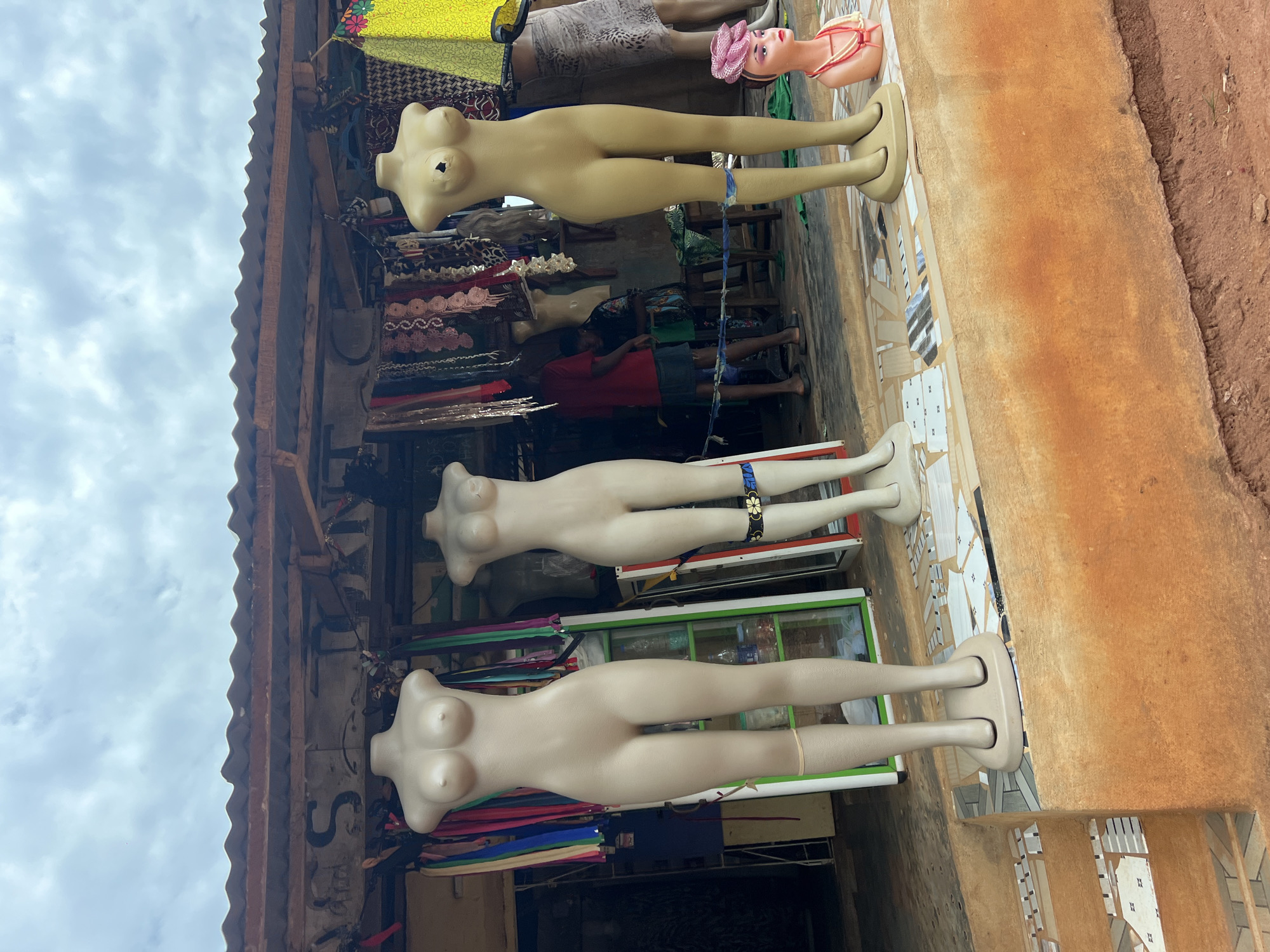 This media file shows a street-side market stall in Nkpor, Nigeria, with mannequins displayed prominently. The scene is outdoors, with a cloudy sky visible overhead. The stall appears to be selling fabrics and clothing, as evidenced by the stacks of colorful textiles in the background and the mannequins themselves, which are torso-only models designed for displaying garments.
Three mannequins are visible. The topmost mannequin is a pale beige color and appears to be older, with a visible hole on its chest. It is adorned with a pink hat and a decorative neckline that appears to be made of beads. Below it, another mannequin, also pale beige, is displayed. This mannequin has a beaded bracelet with floral accents on its arm. The third mannequin, positioned at the bottom, is a lighter, almost white color, and is presented without any adornments.
The mannequins are arranged vertically, suggesting they are leaning against the stall structure or a display rack. The background reveals more of the market, including shelves stocked with various fabrics, some folded and others rolled. A person wearing a red shirt and blue shorts is partially visible behind the second mannequin, seemingly engaged in market activities. The ground in front of the stall is paved with mosaic tiles. To the right of the stall, a dirt path and a wall made of reddish-brown earth are visible. The overall impression is one of a bustling marketplace, likely in the daytime given the natural lighting. No visible text is present in the image.