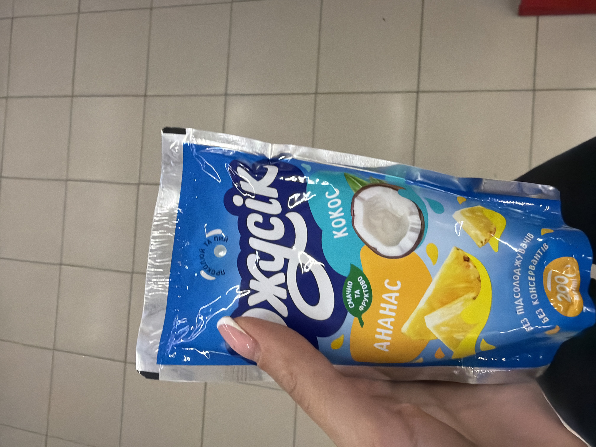A hand with a french manicure is holding a blue pouch of juice with images of pineapple and coconut. The juice pouch has text in Ukrainian. The background is a light-colored tiled floor. The scene appears to be indoors, possibly in a grocery store or similar retail environment. No specific location in Kyiv, Ukraine can be determined from the image.