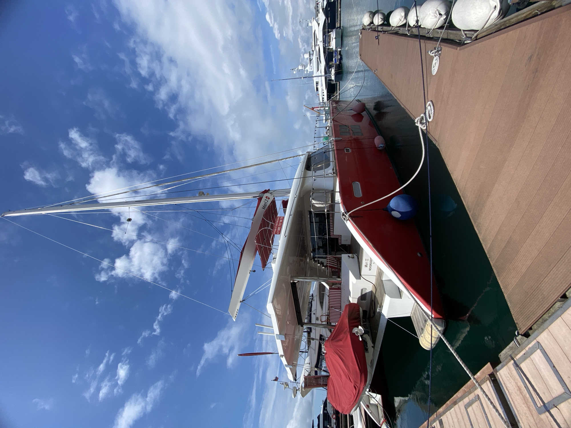 The image depicts a red and white catamaran sailboat docked at a pier in Sochi, Russia. The sailboat is moored to the pier with ropes. The sky is blue with scattered white clouds. A series of white spherical fenders are attached to the pier. Other yachts are visible in the background. A red protective cover is on the stern of the boat. The name "Meri II NOVOROSSIYSK" is visible on the boat. The pier is constructed of brown planks.